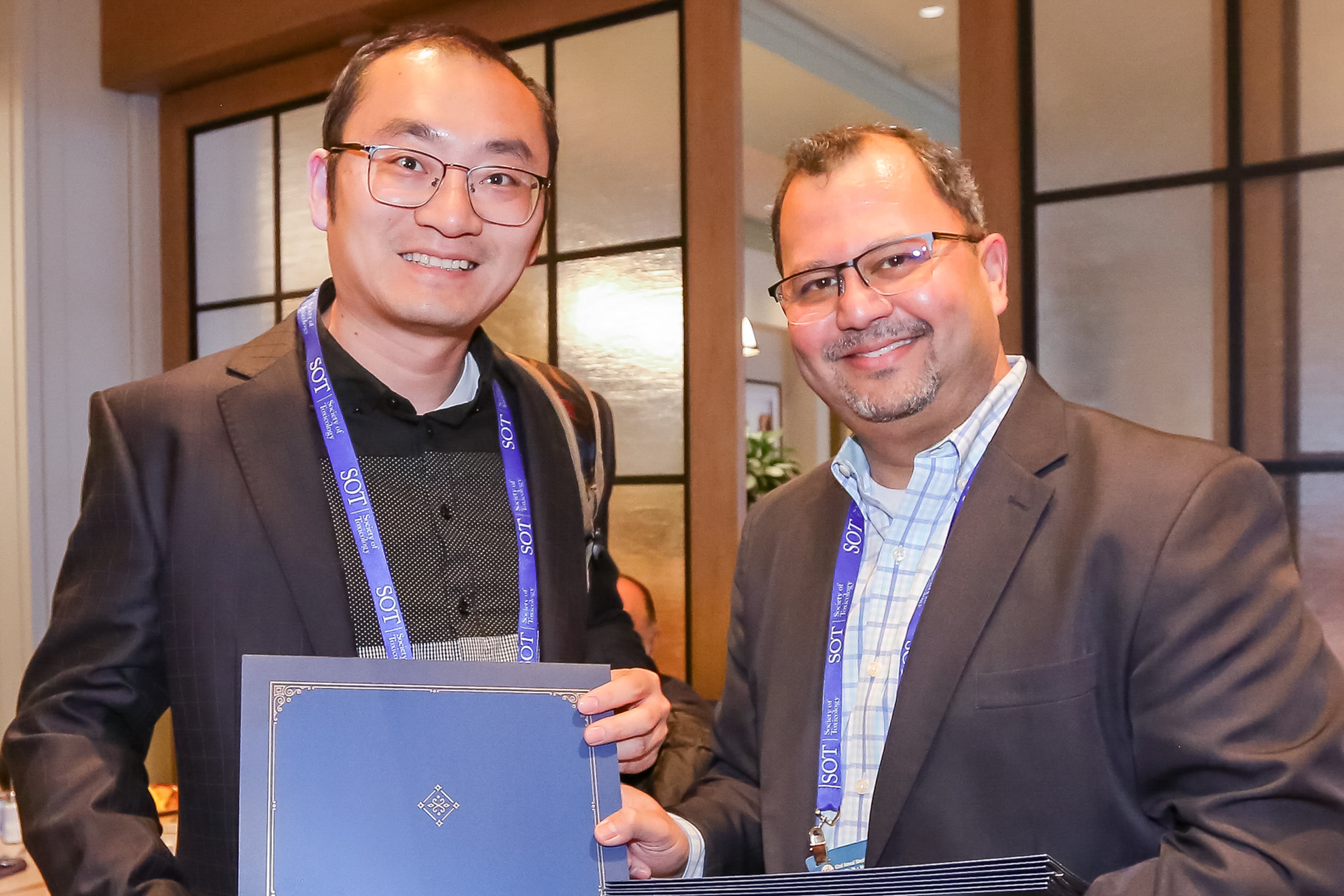 Two men are standing beside each other and smiling at the camera. Both are coming together to hold an award. The man on the left has a stack of awards in his hand.