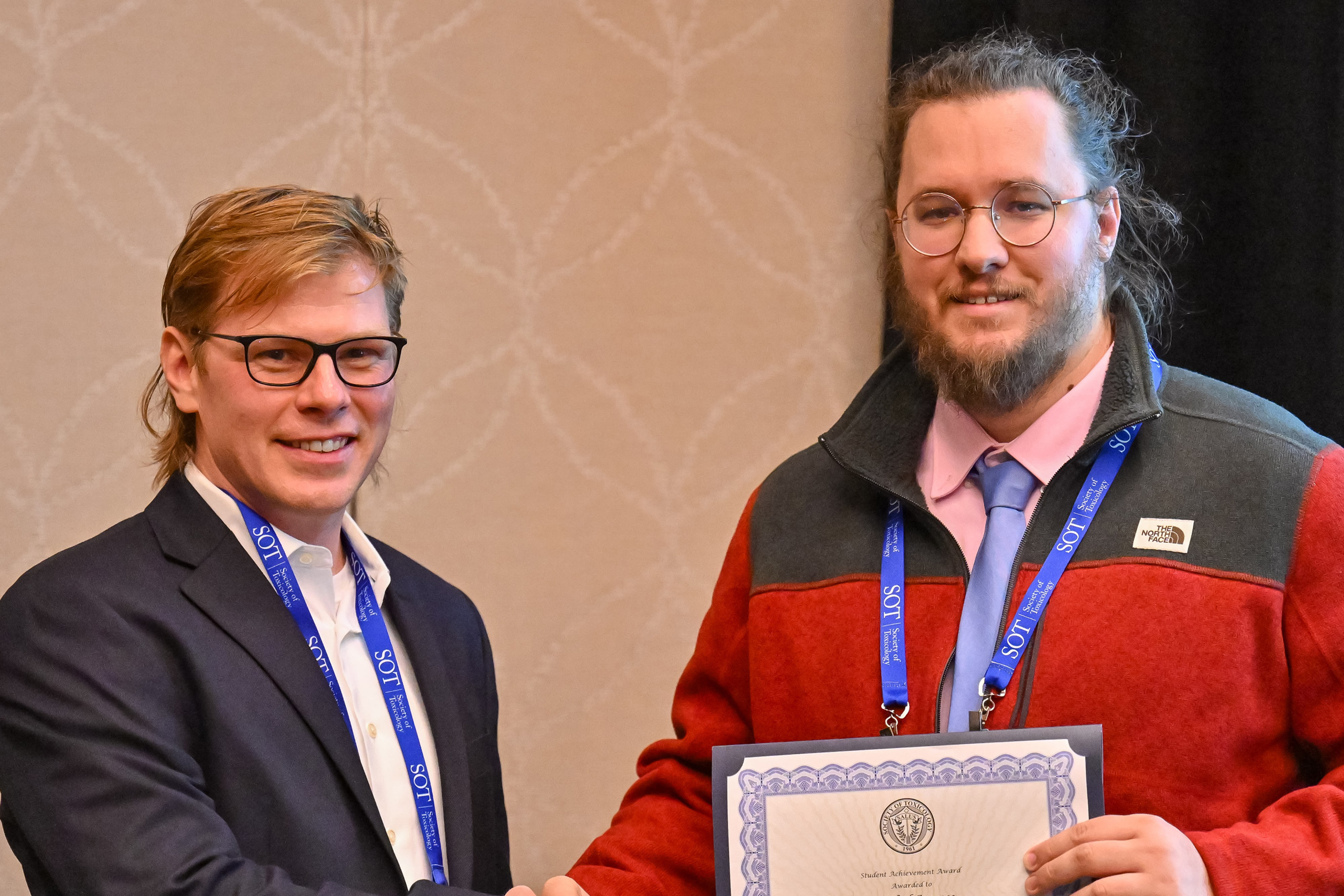 Two men are standing beside each other and smiling at the camera. The man on the left is shaking the other man's hand. The man on the right is holding an award plaque in his left hand.