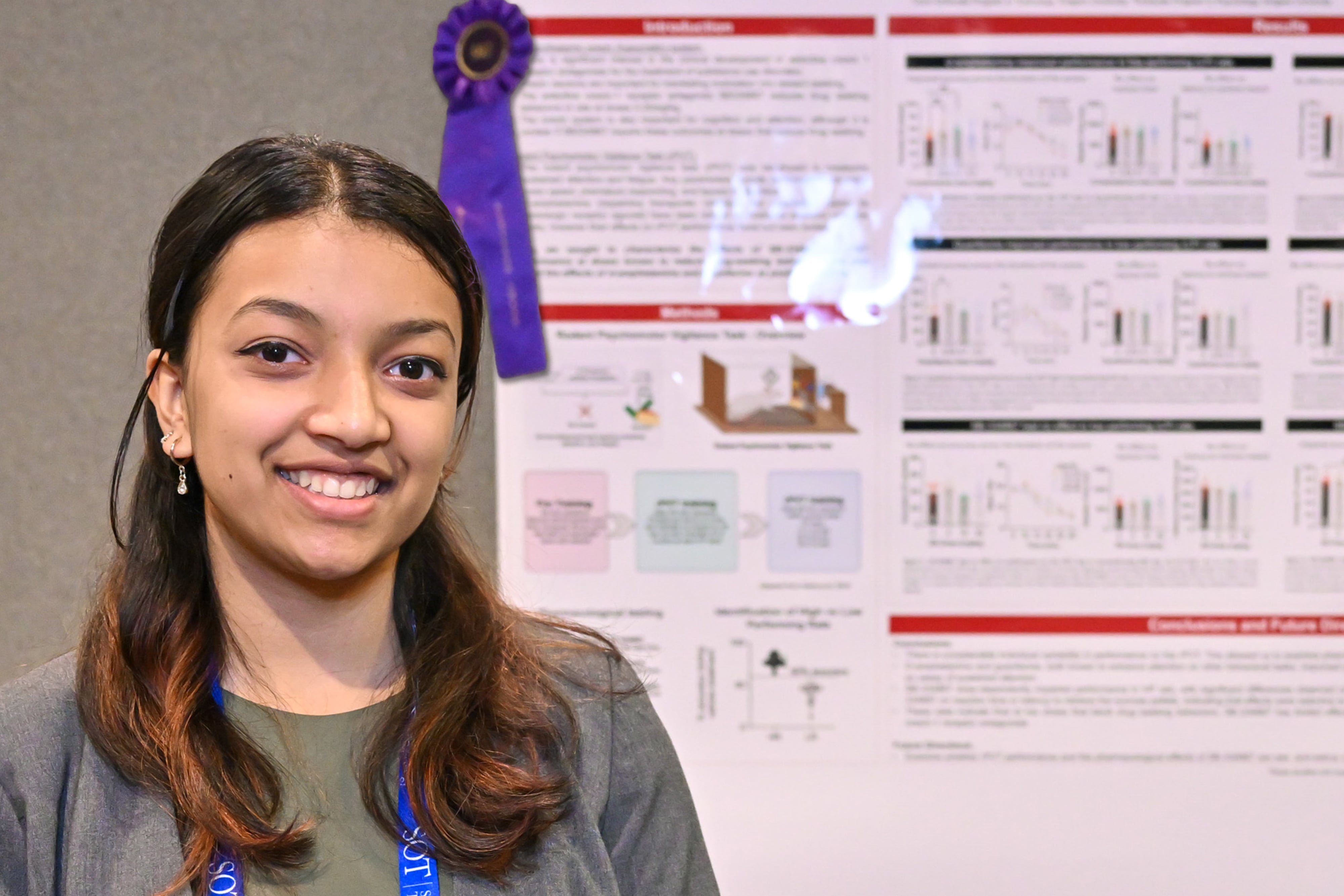 A woman is standing to the left of a poster board, smiling at the camera. The poster board has a blue award ribbon on it.