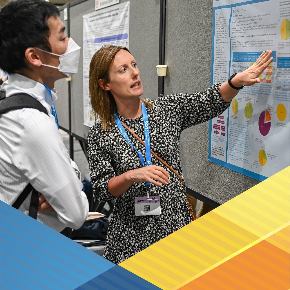 In the center of the image is a woman gesturing to her left at a poster hanging on a gray poster stand. She is looking at the poster while talking to a man standing to her right who also is looking at the poster.