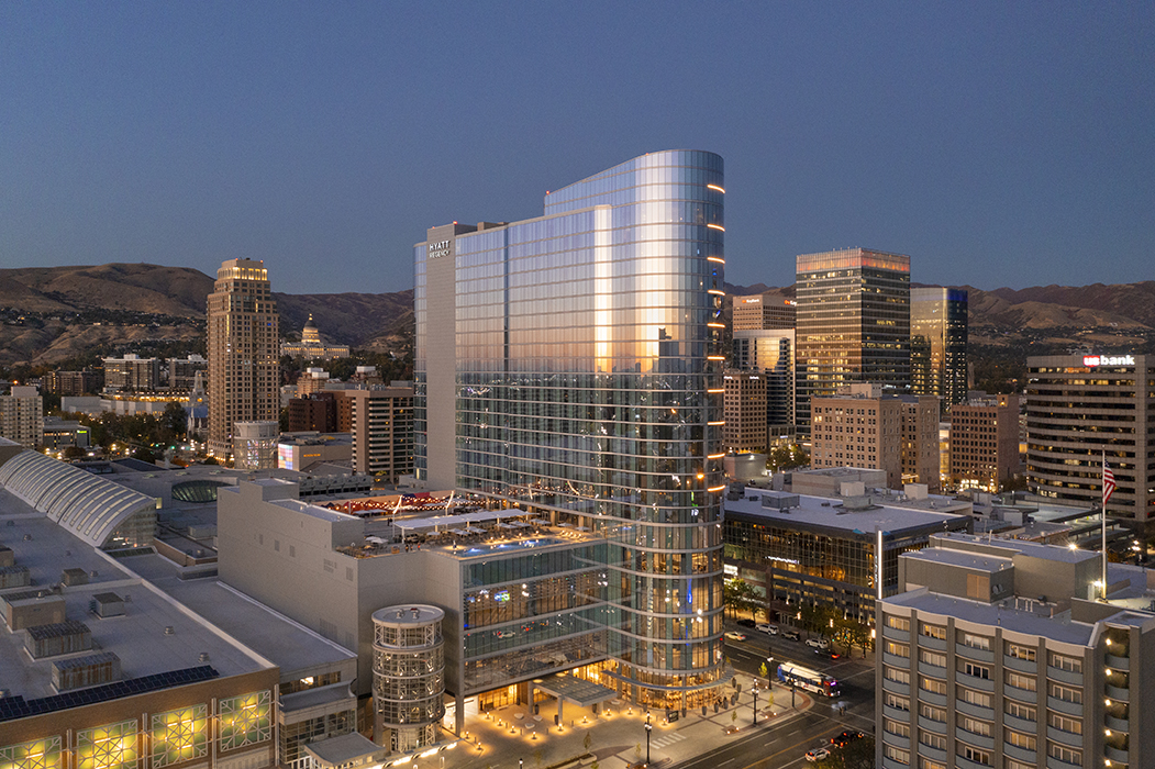 It is twilight in downtown Salt Lake City. Lit in the center is the Hyatt Regency, which consists of a tall, multi-story building with large glass windows and a shorter connected multi-story addition with a pool and lounge area on the top. Surrounding the Hyatt Regency are other Salt Lake City buildings.