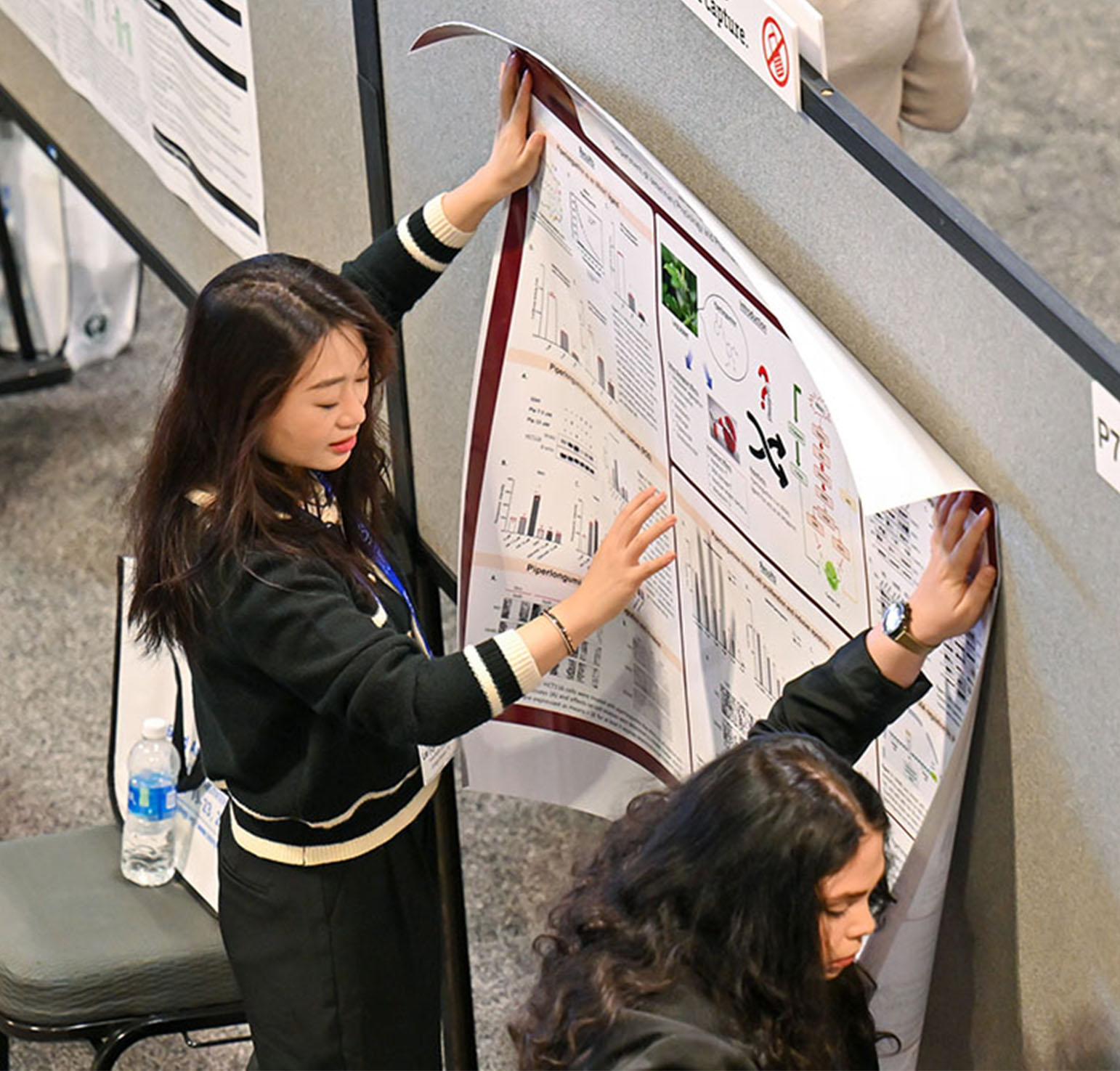 An overhead shot shows two individuals holding a large poster against a gray fabric-covered board. They appear to be in the process of attaching the poster to the board.