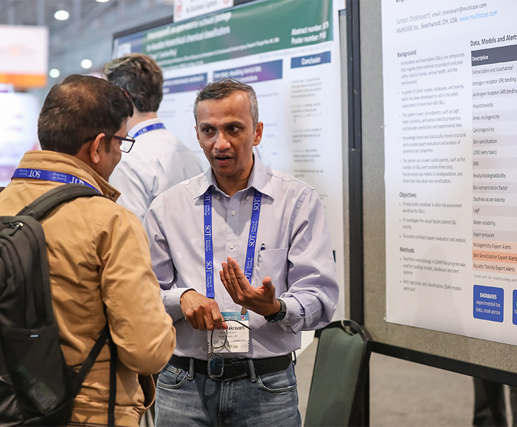 An individual is standing near a poster display. This individual is speaking with another individual and appears to be explaining something based on the facial expression and position of the hands.