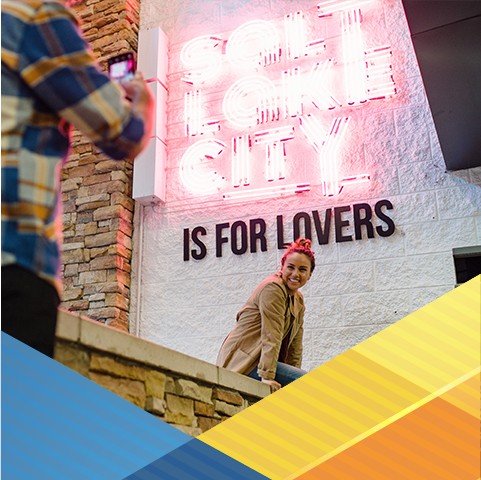 A young individual is sitting on a brick wall, turned sideways toward an individual taking a picture. On the wall next to the seated individual is a pink neon sign reading Salt Lake City. Below the neon sign in black, 3D lettering is the phrase Is for Lovers.