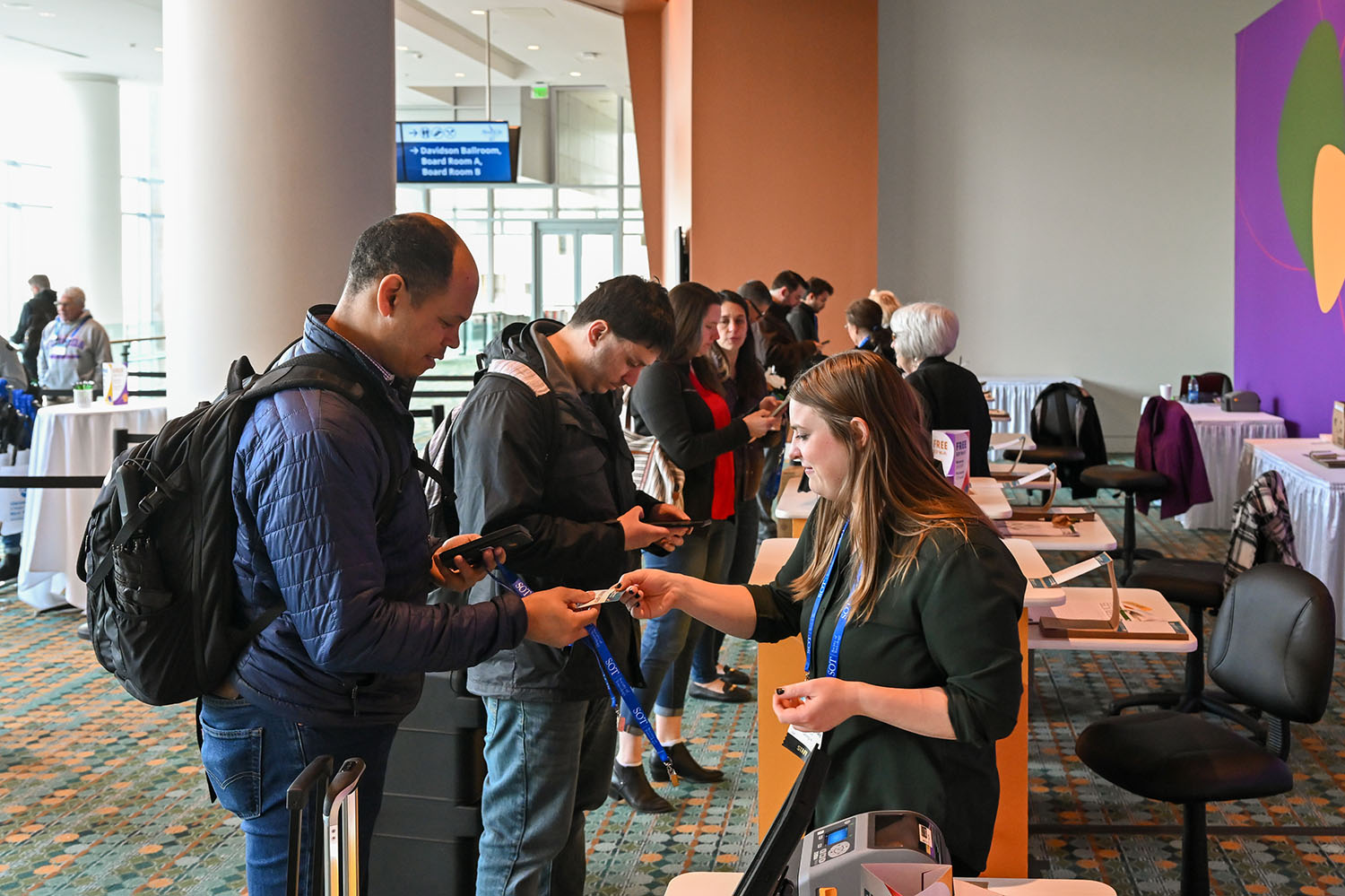 A man with a backpack over his shoulder is receiving a paper badge from a woman standing in front of him. Other individuals are seen in the background standing at registration kiosks.