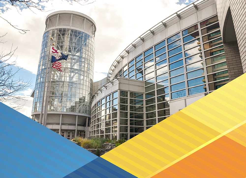 This image is an exterior shot of the East Tower of the Salt Palace Convention Center. The tower is multi-stories tall and is all glass. It is connected to a building with a curved wall that also is all glass.