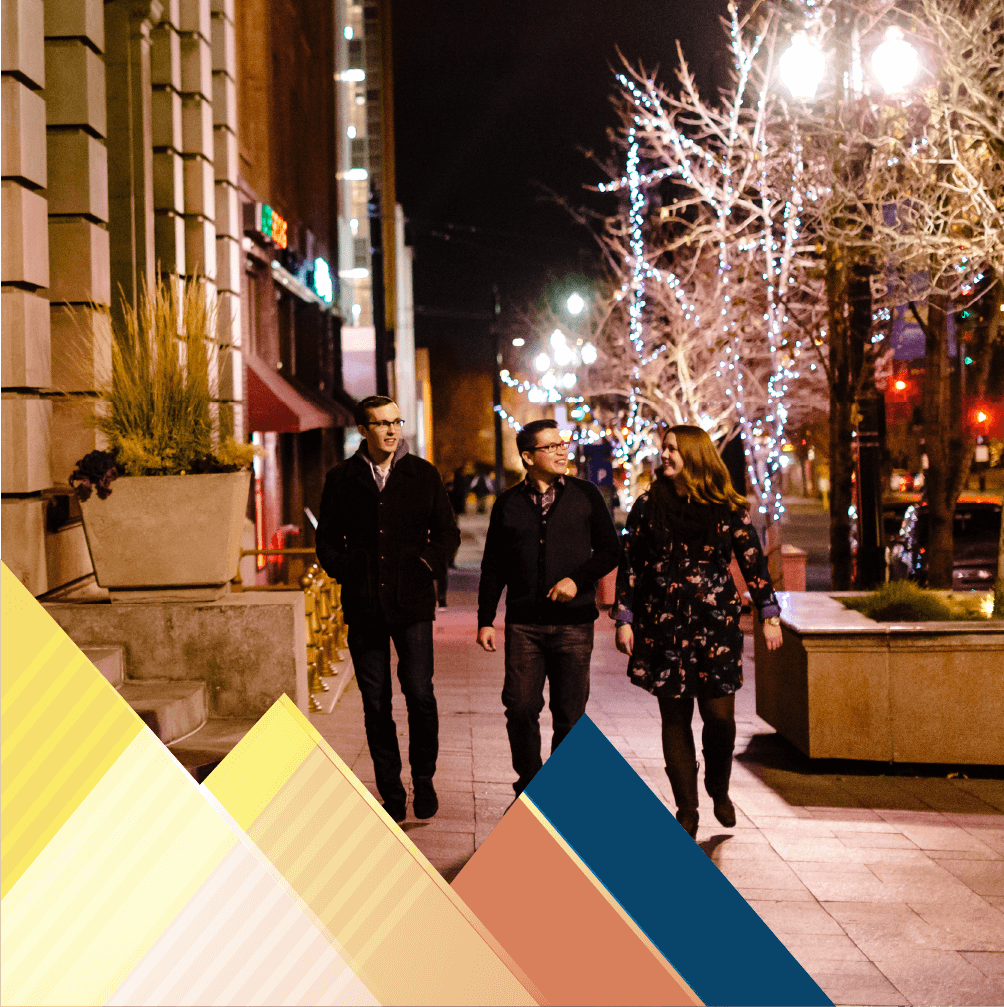 Three individuals are walking down a sidewalk toward the camera on a city sidewalk at night.