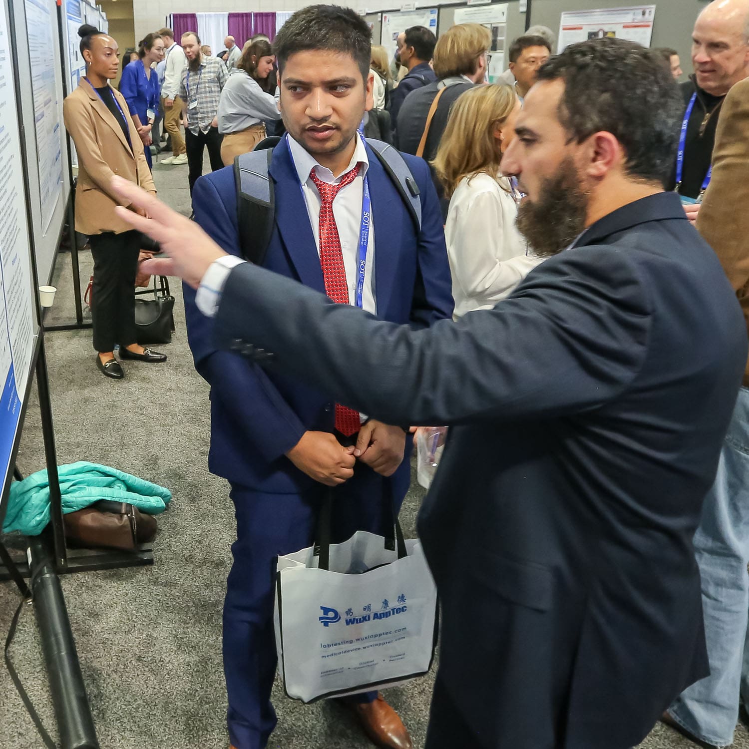 Two individuals stand looking at a large poster display that is on the left-hand side of the photo. One individual is gesturing to the poster. Many additional poster displays and individuals in conversation can be seen behind the two individuals.
