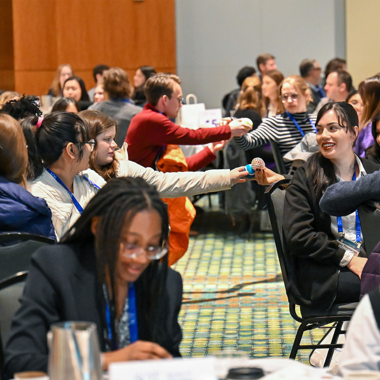 The image depicts a room full of young people. They are seated in chairs with a large aisleway between them. Individuals on one side of the aisle are passing different kinds of balls to individuals on the other side of the aisle.