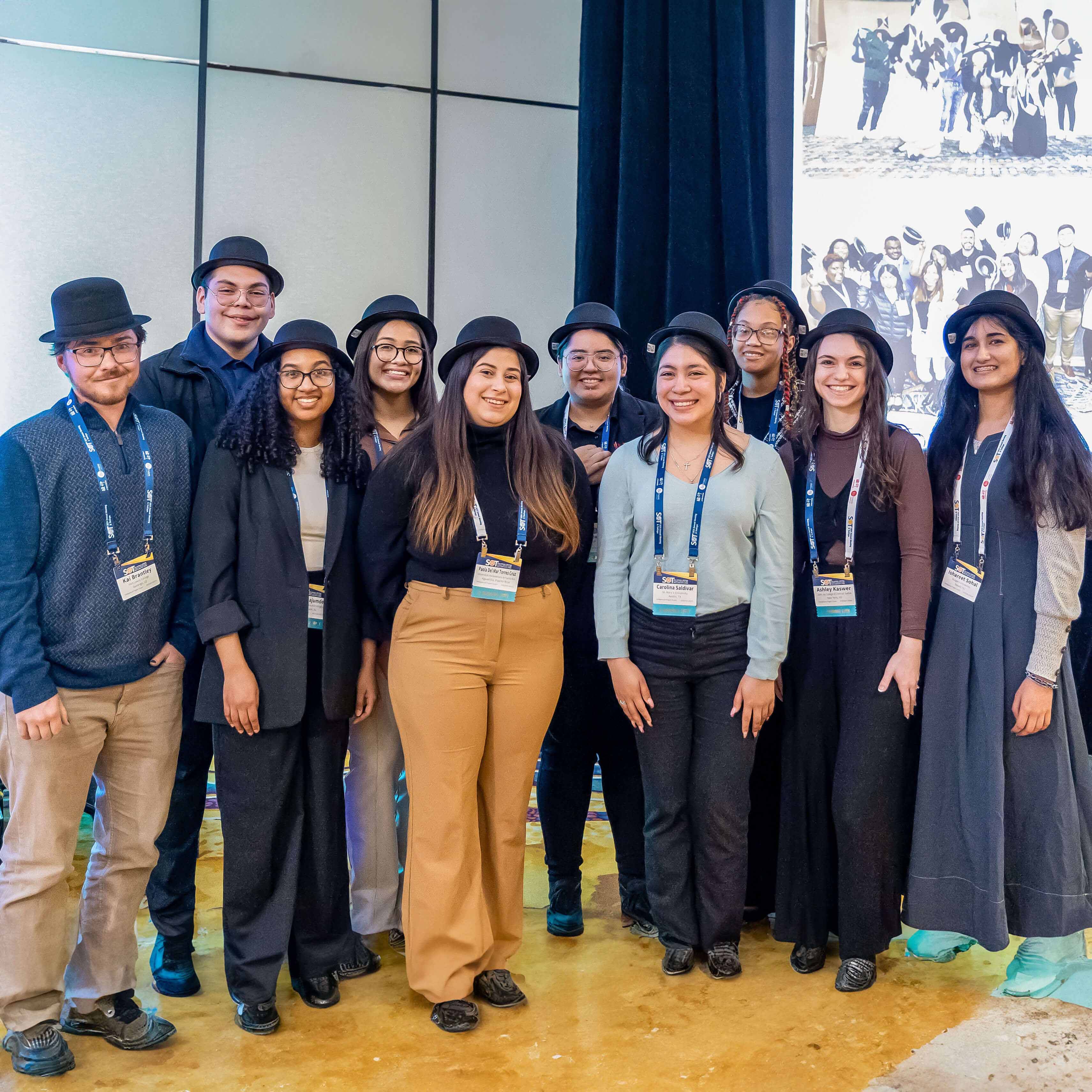A diverse group of young individuals are posed for the camera. All the individuals are wearing black bowler hats and are smiling.