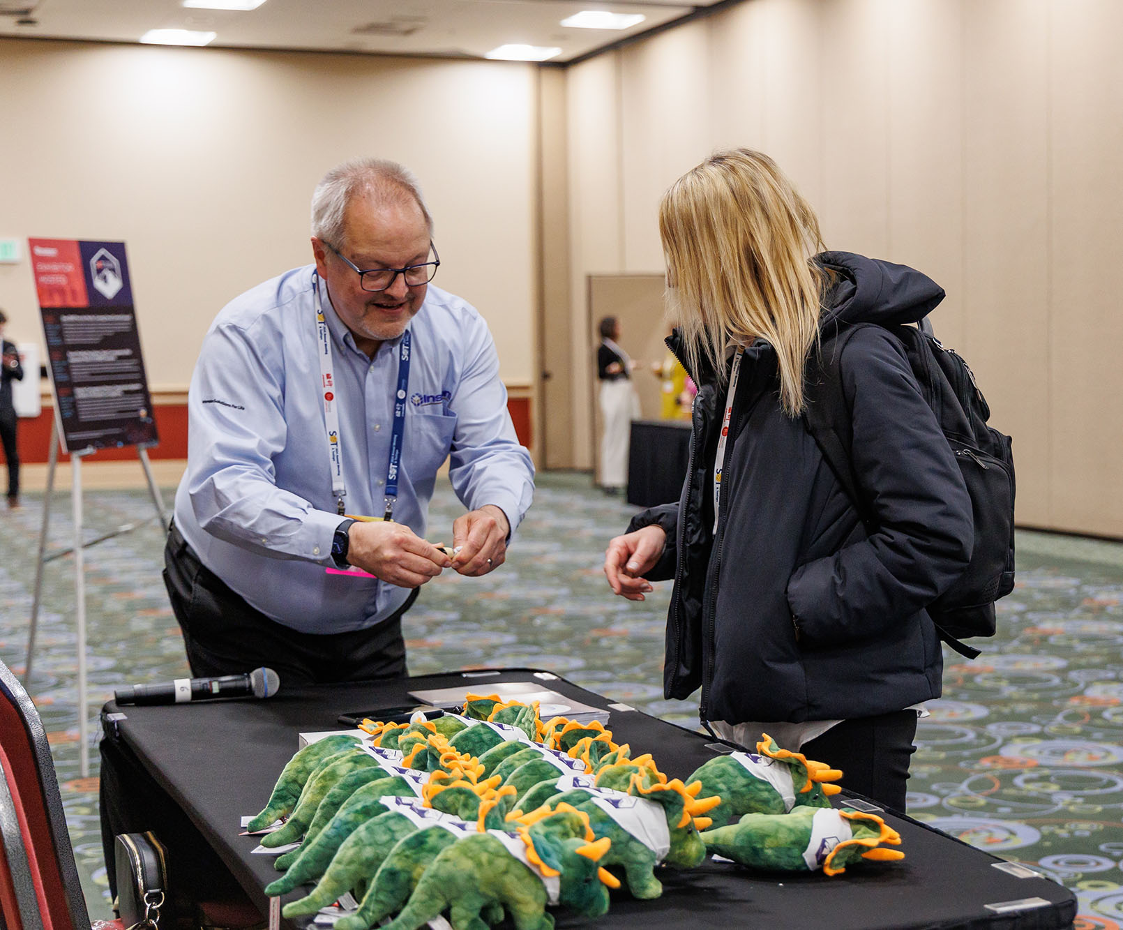 Two individuals stand leaning over a table. One individual is showing the other individual something in their hands. On the table are plush dinosaurs.