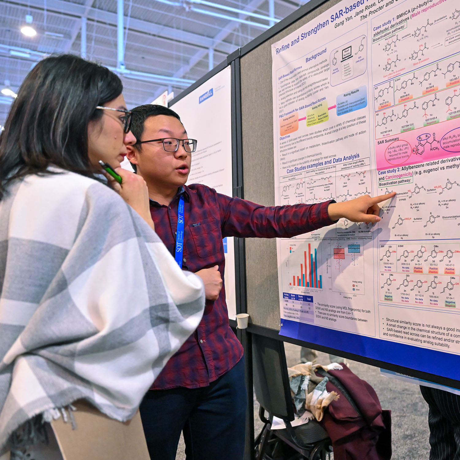 Two individuals stand near each other. Both are looking at a poster hung on a large gray freestanding tack board. One of the individuals is pointing at a figure on the poster and speaking while the other individual is intently listening.
