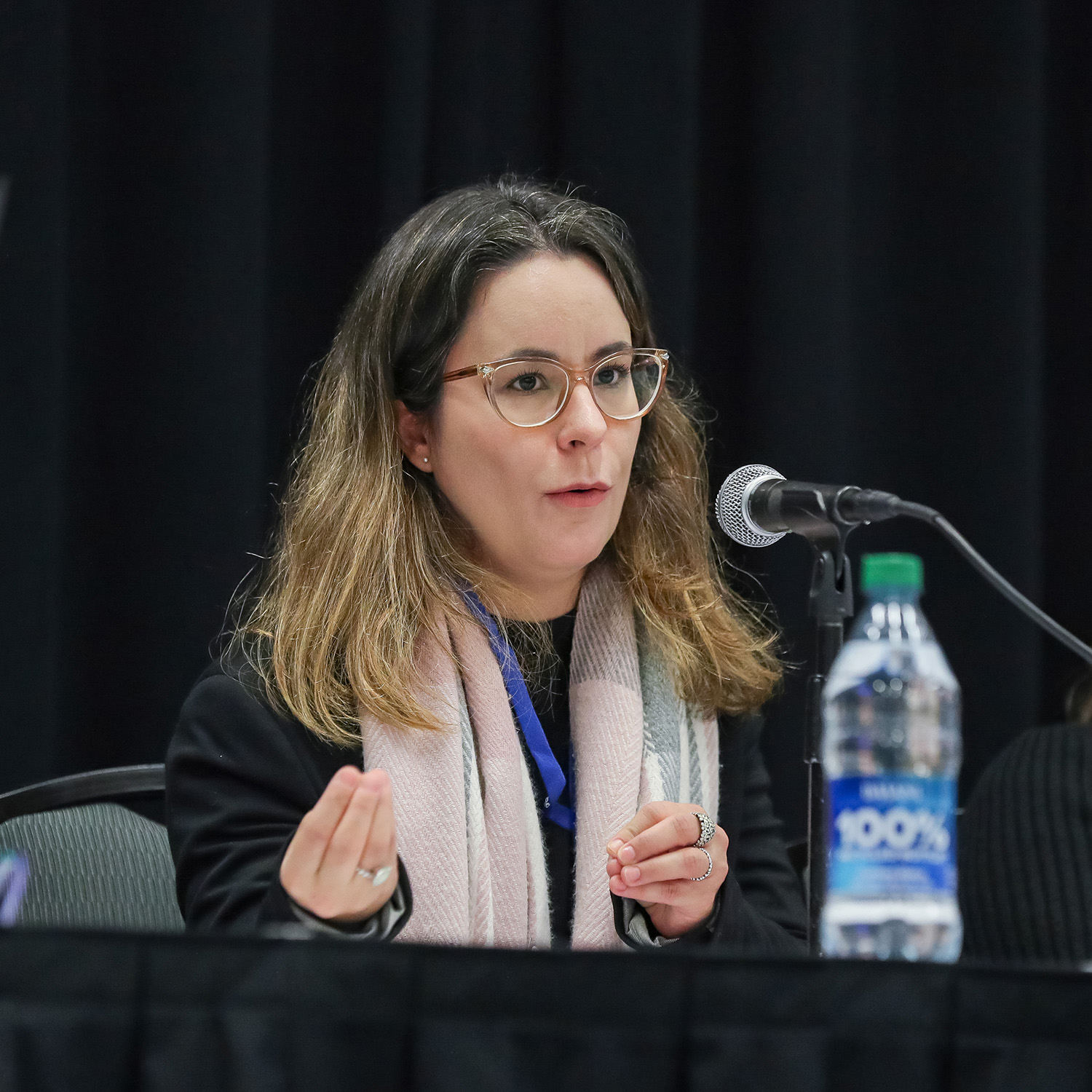 An individual is seated at a table. There is a microphone on a stand sitting on the table in front of them. They are speaking into the microphone while gesturing with their hands.