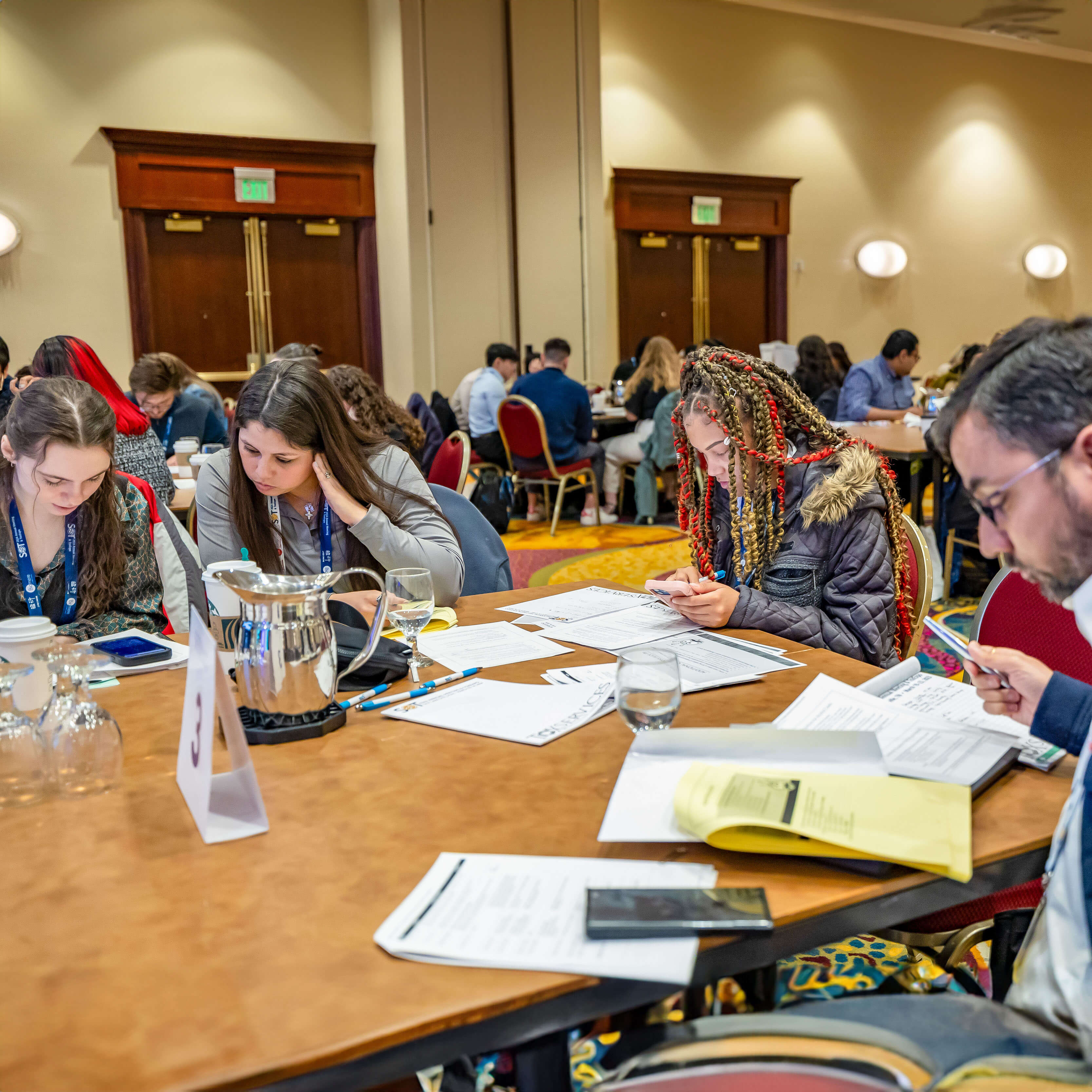 Young people sit at a table. They are focused on papers in front of them and appear to be doing calculations using their phone.