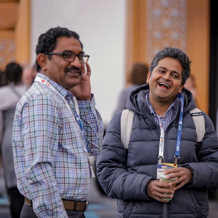 Two individuals are standing near each other socializing. They are smiling and looking off camera.
