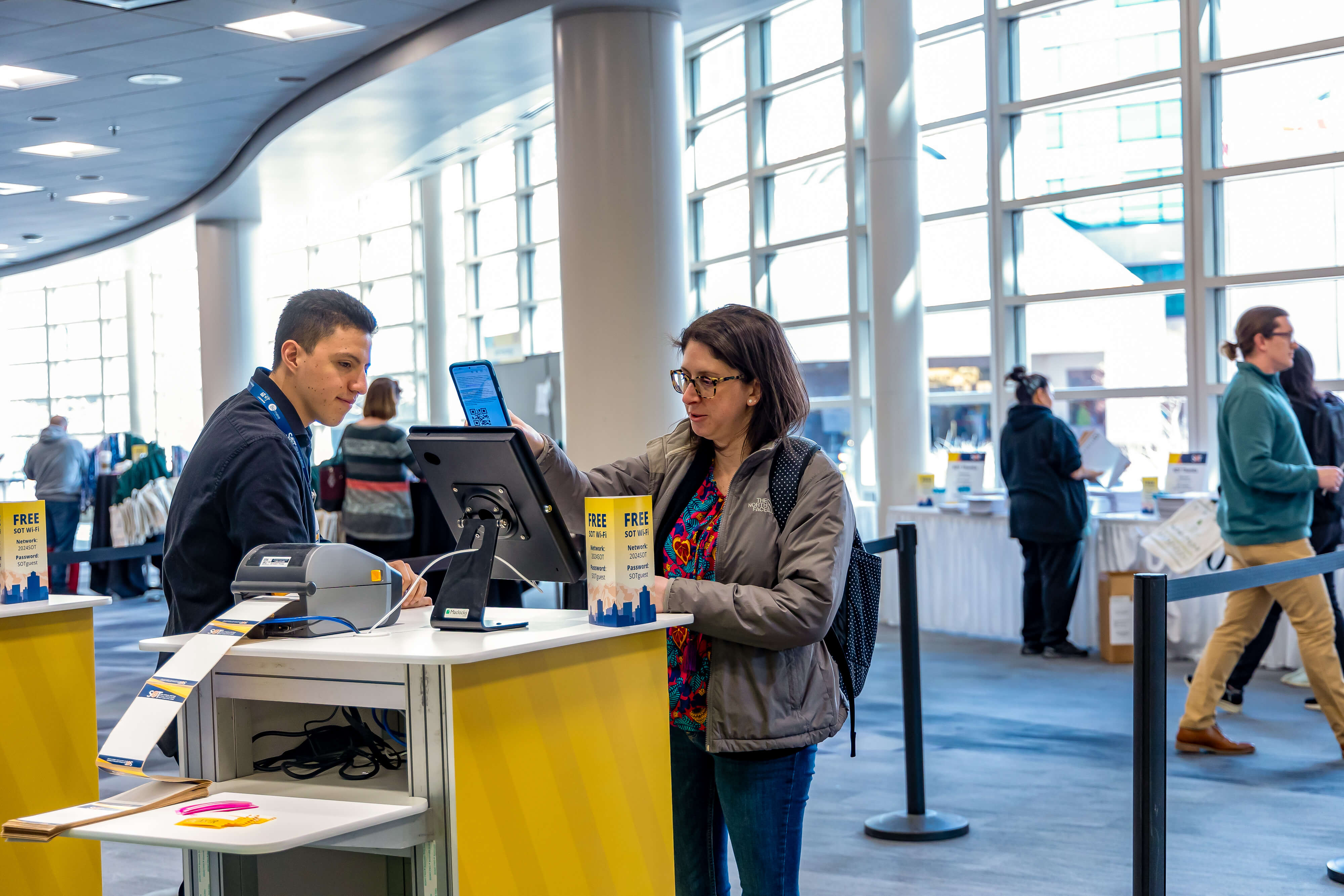 An individual stands in front of a kiosk. On top of the kiosk is a mounted screen. The individual is holding their phone in front of the screen to scan a QR code. A registration assistance is standing next to the kiosk in case help is needed.