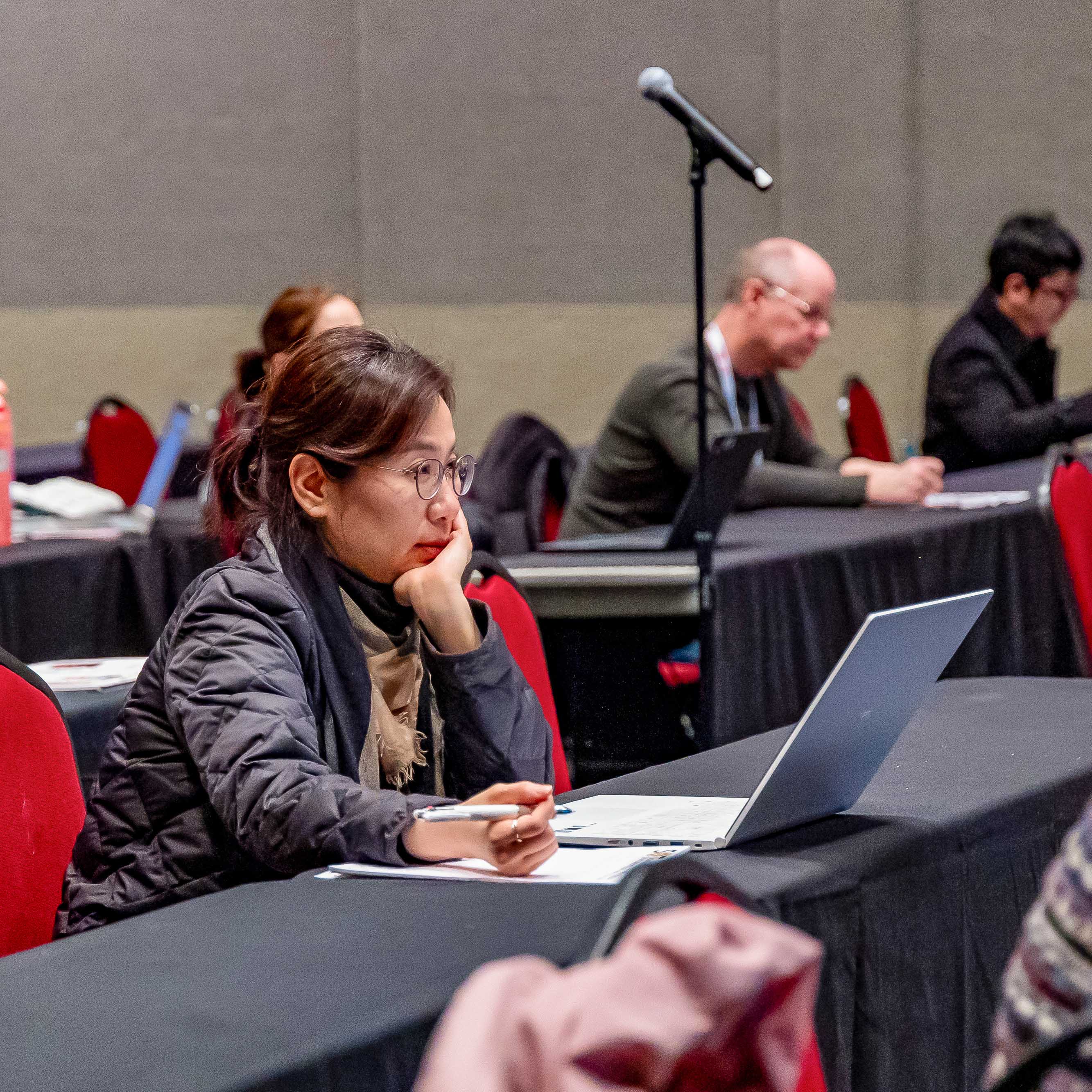 An individual sits at a long table. A laptop sits open on the table. The individual is holding a pen, ready to take notes on a notepad. They are looking toward the front of the room in concentration.
