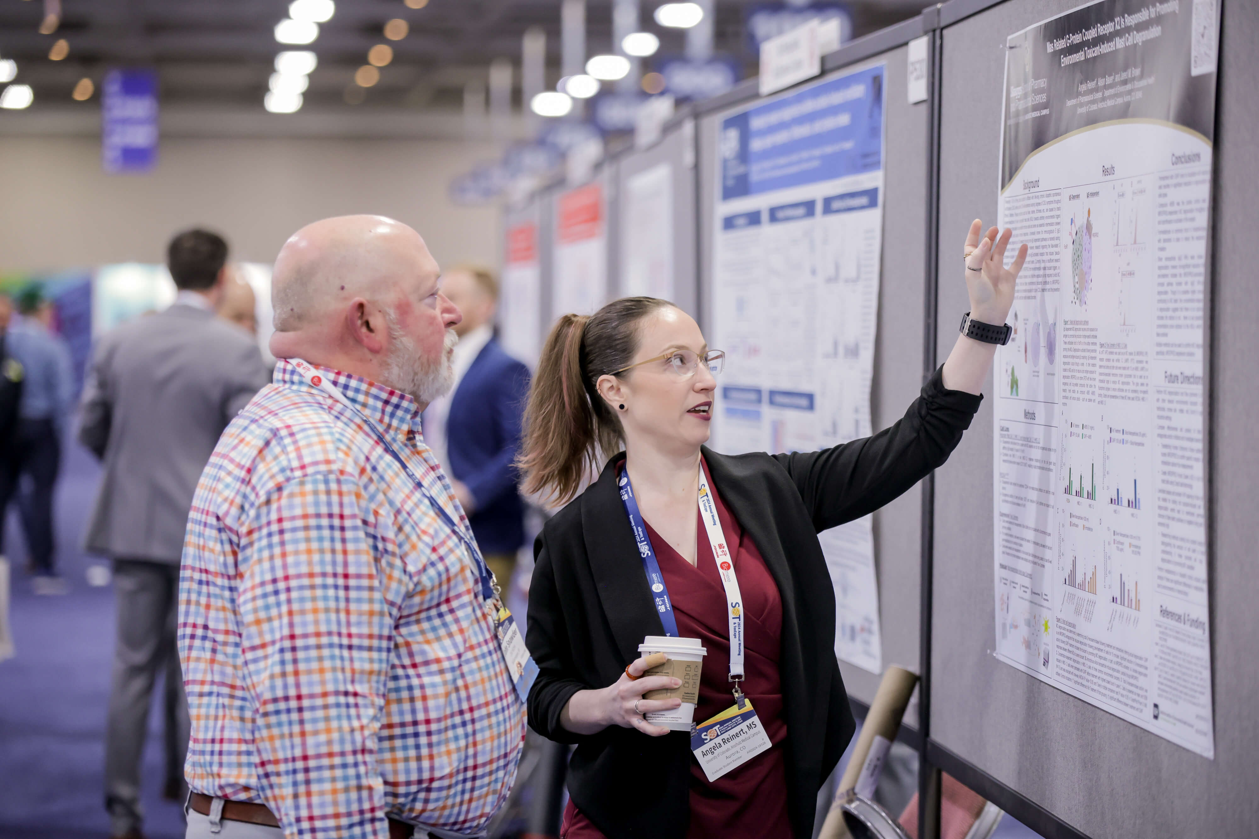 Two individuals stand next to a large gray board that holds a scientific poster. Both are looking at the poster. The individual closest to the poster is speaking and gesturing at the poster with one hand.