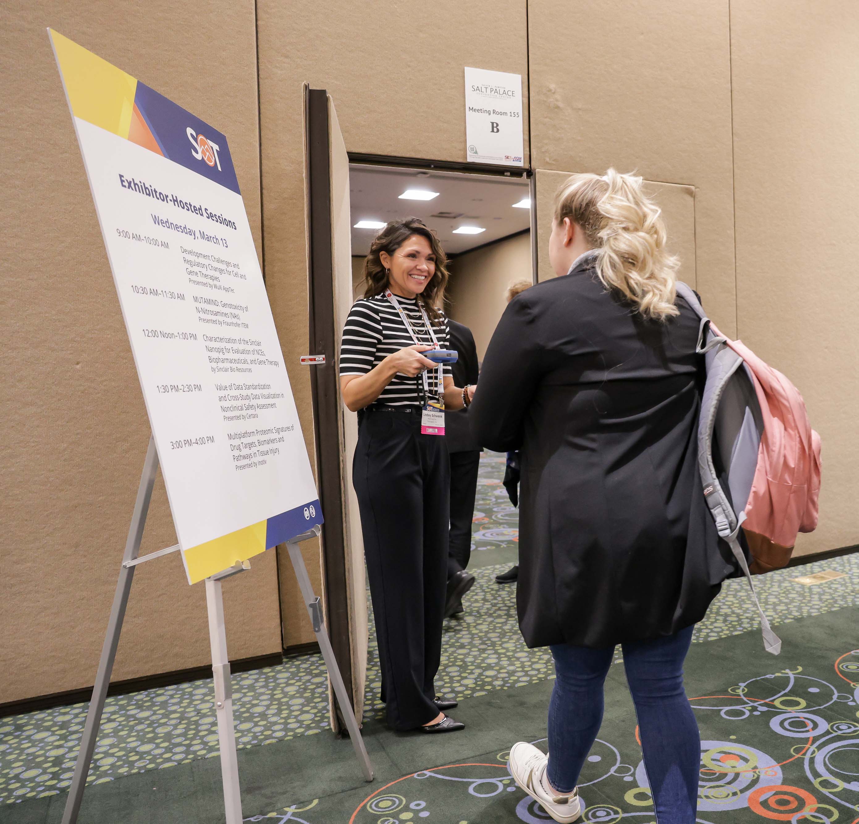 A smiling individual stands in front of a door holding a scanning device. Two individuals are standing in light in front of the individual at the door, waiting to be scanned in order to enter the room.