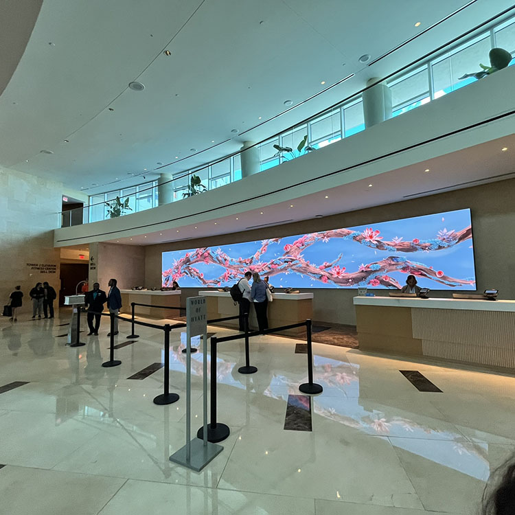 An interior view of a hotel lobby. The flooring, walls, and ceiling are light and bright. Behind the check-in desks is a large digital display showing a branch with flowers.