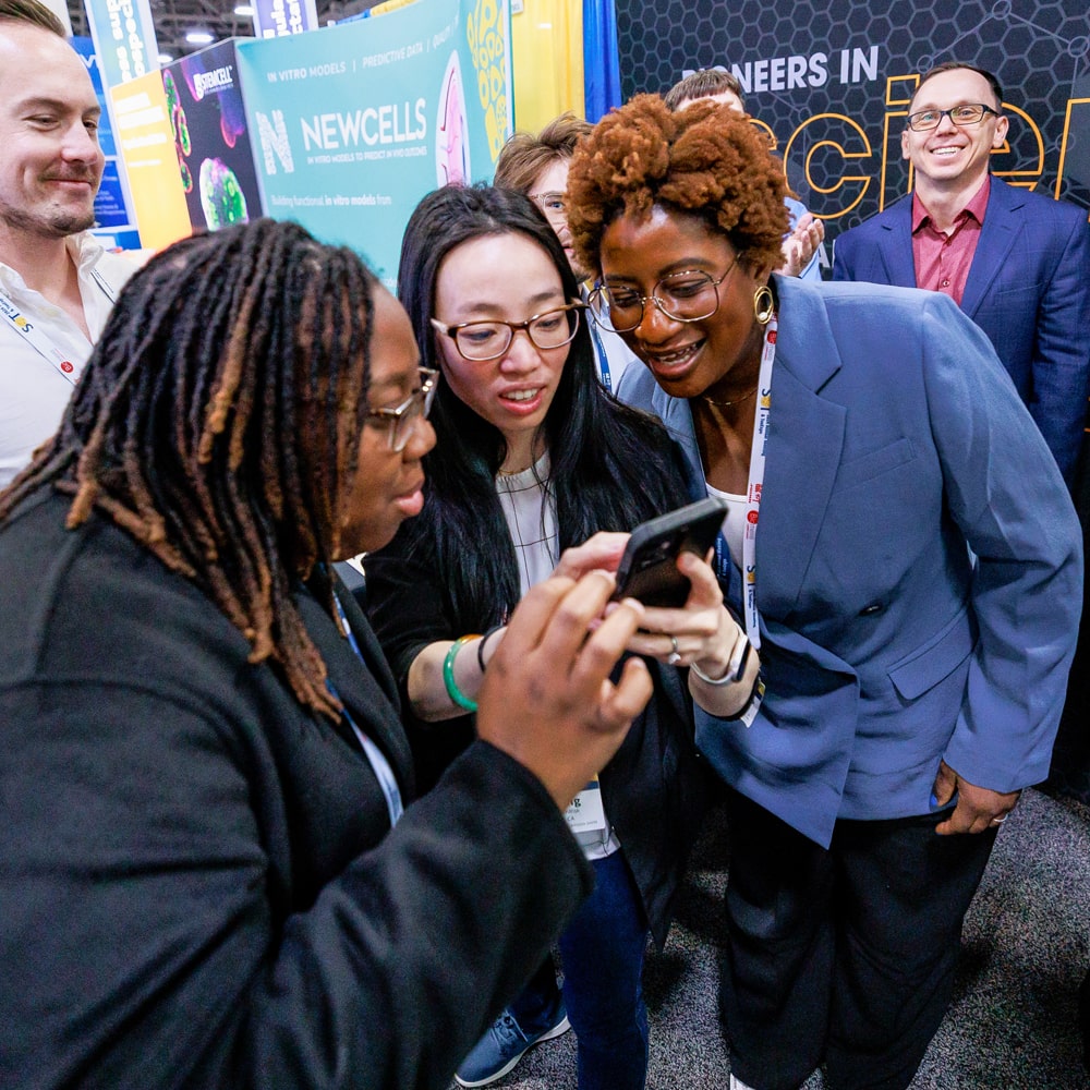 Three individuals stand crowded together with other individuals around them. The three individuals are looking at a phone held by the person in the middle of the small group.