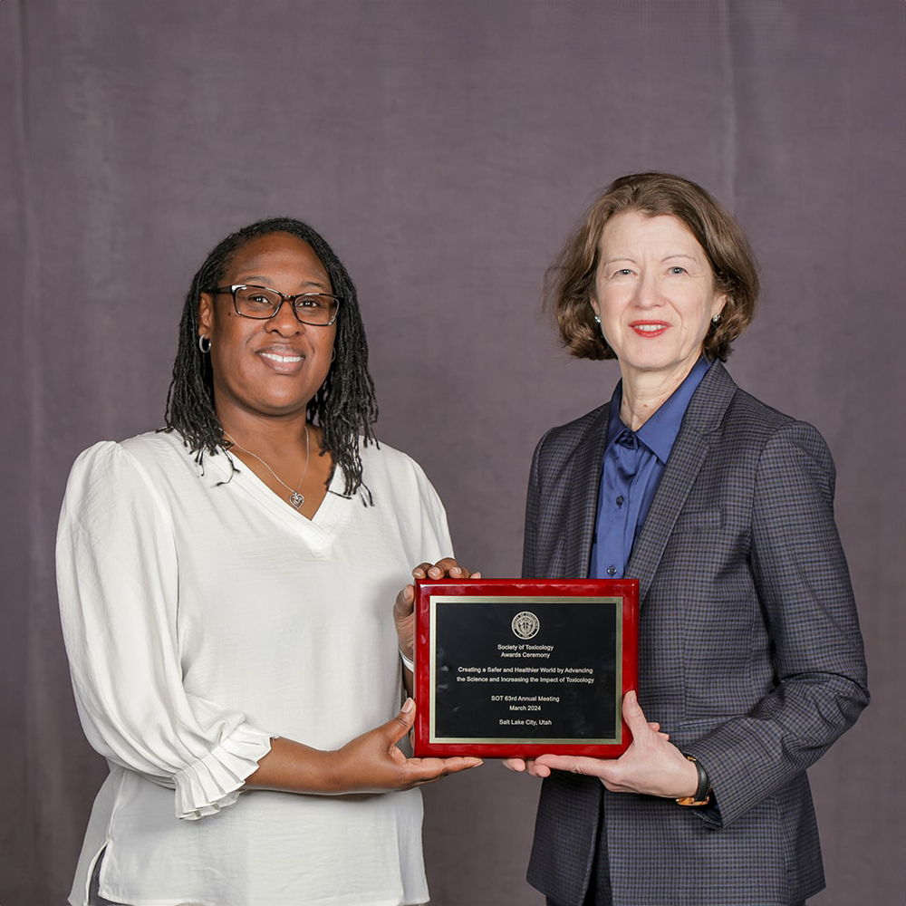 Two individuals are smiling at the camera and holding an award plaque between them.