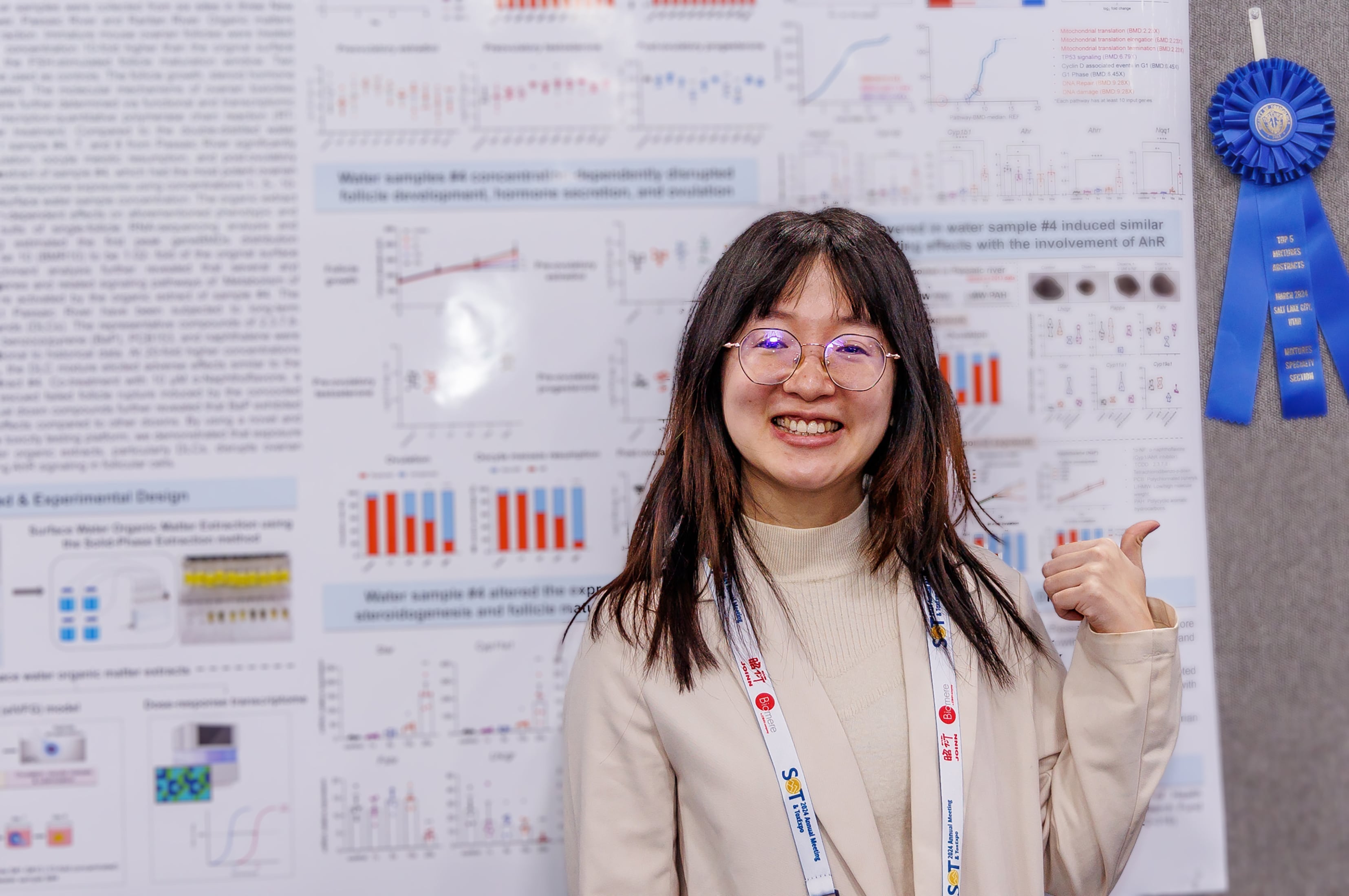 An individual stands in front of a mounted poster board. They are smiling at the camera and using their thumb to point behind them at the blue ribbon tacked next to the poster.