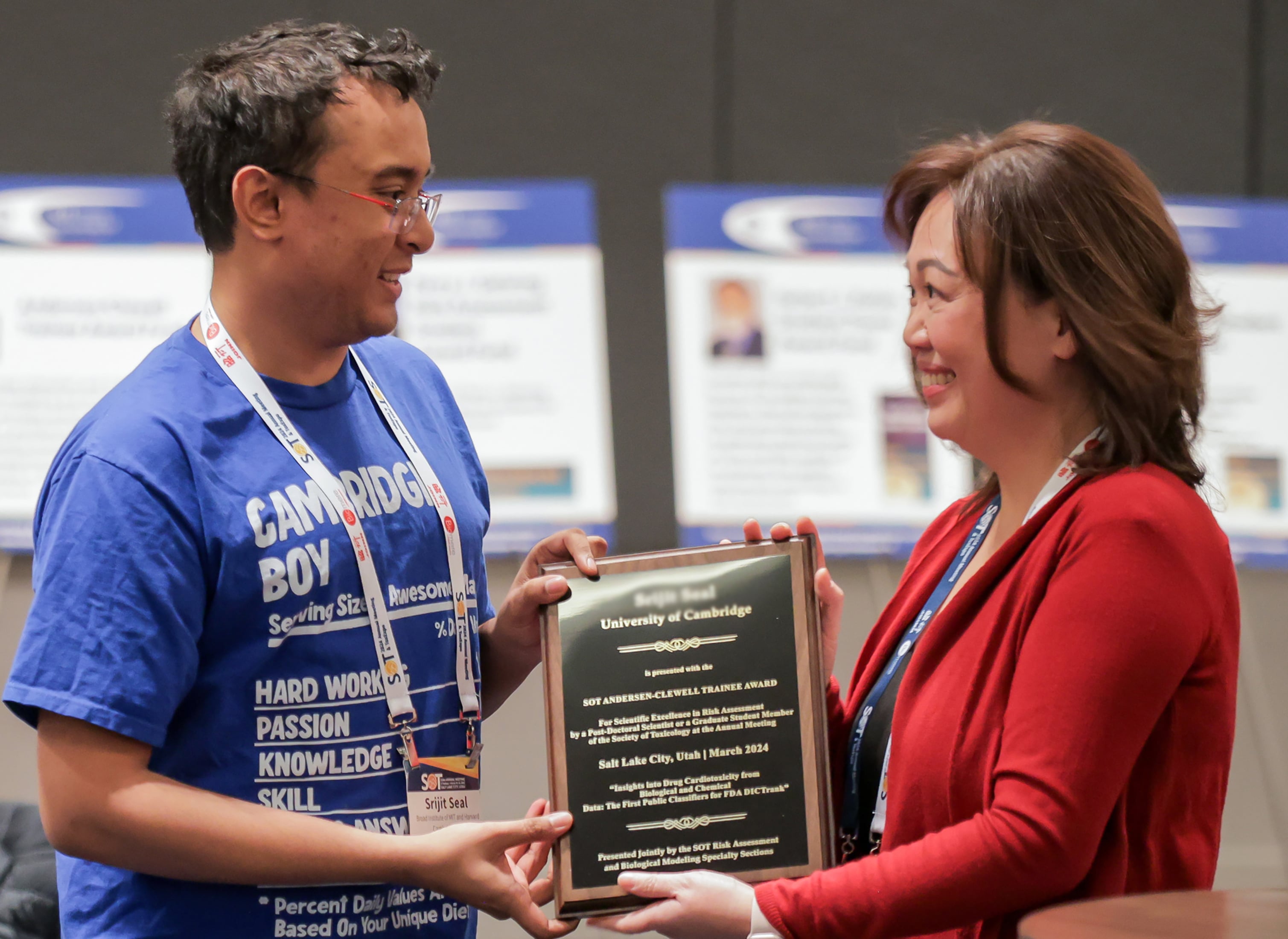 Two individuals are smiling at each other. One is handing a large award plaque to the other. Surrounding the individuals are round tables where people are seated.