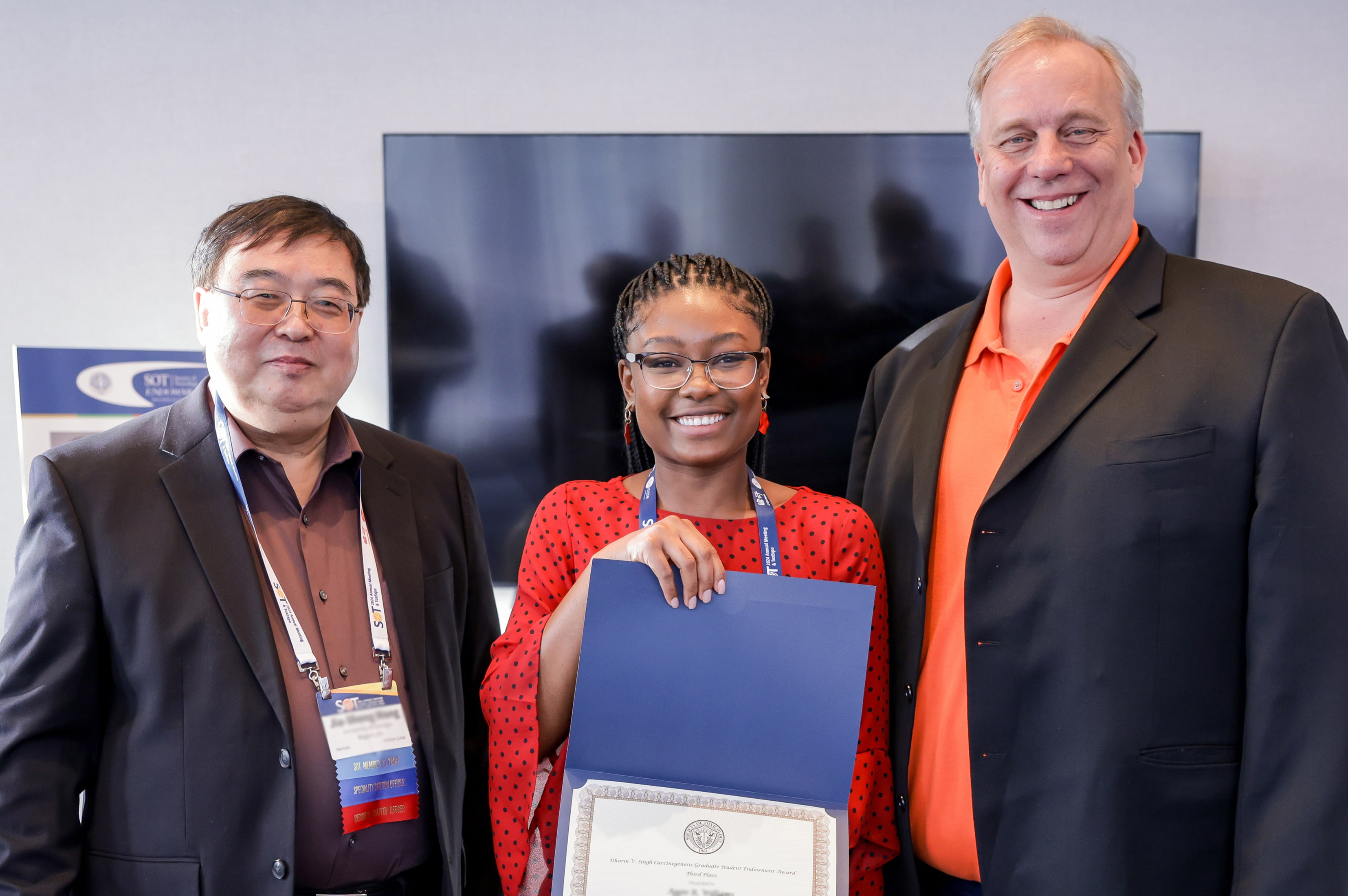 Three individuals stand next to each other and are smiling at the camera. The individual in the middle is holding an award certificate.