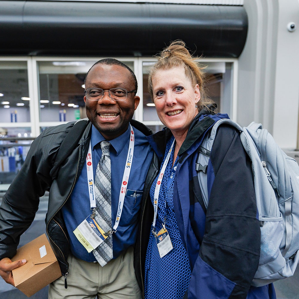 Two individuals stand next to each other with one arm around the other person’s back. They are smiling at the camera.