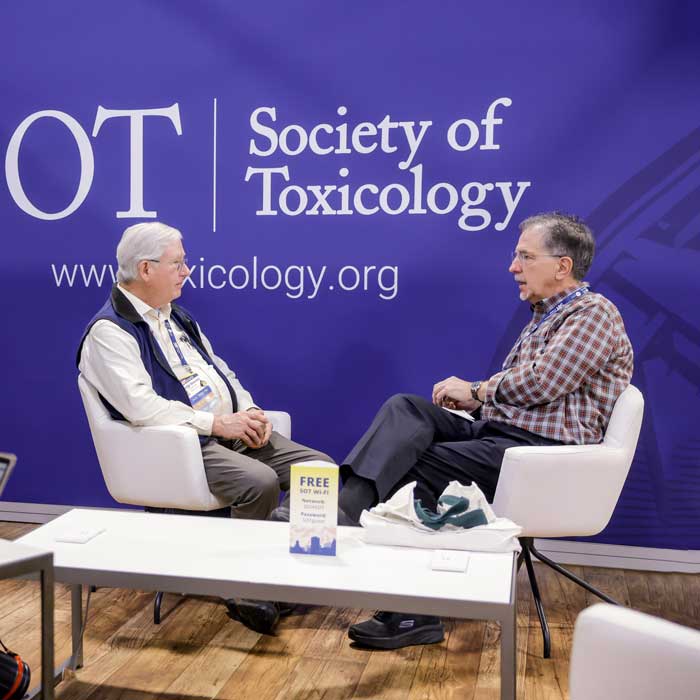 Two individuals are sitting in swivel chairs next to a coffee table and in the midst of a discussion. They are seated in front of a blue wall that contains the SOT logo.