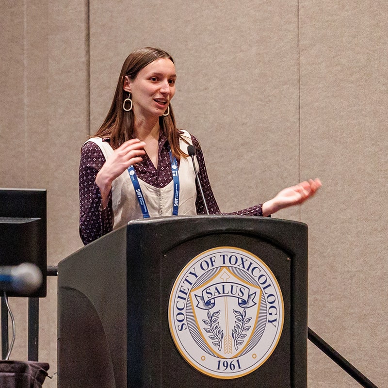 An individual stands behind a lectern that has the SOT salus on its front. The individual is speaking and gesturing.