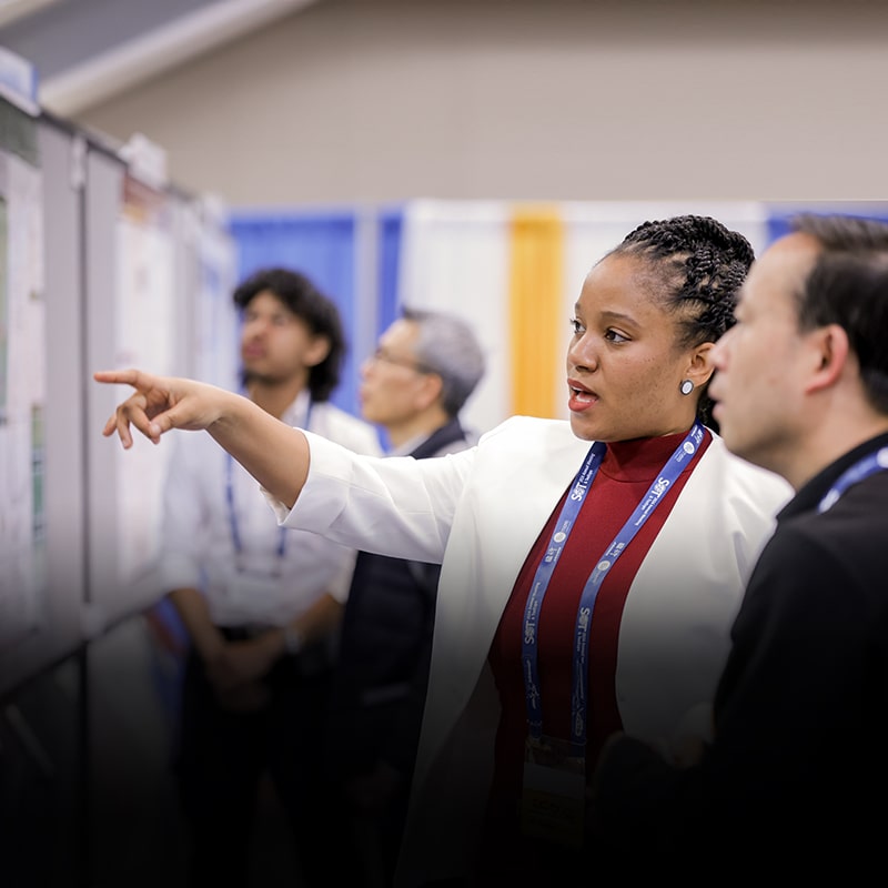 An individual is in profile pointing toward a poster board. Another individual stands next to them, listening to what they are saying. You can see other posters and individuals in the background.