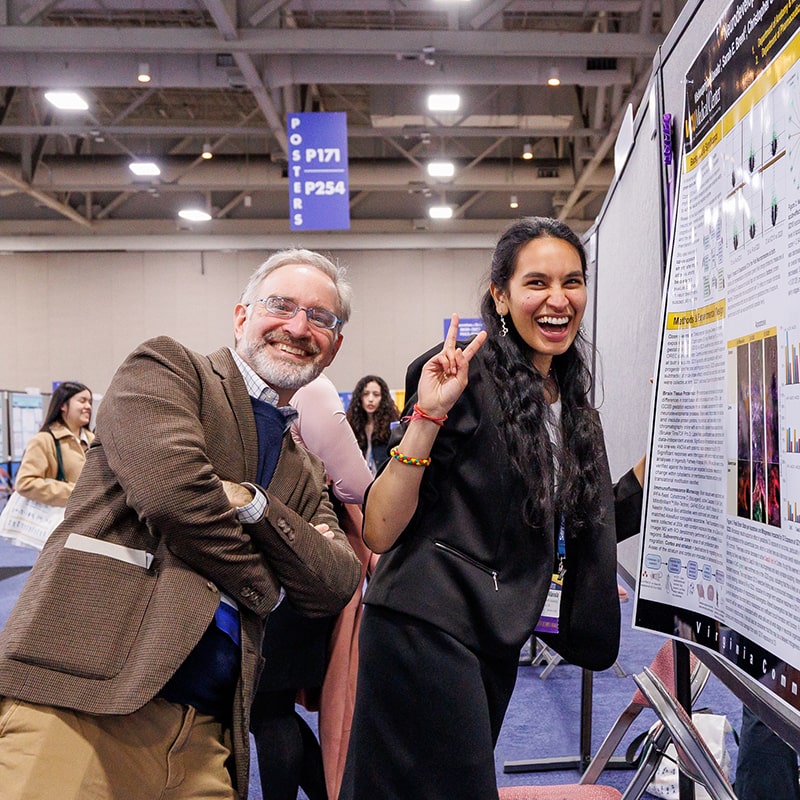 Two individuals are smiling at the camera, with one holding up two fingers in a “V” shape. They are standing next to a poster board with a poster mounted on it except for one final corner. One of the individuals has their hand under the corner; they are clearly getting ready to place the final push pin into the corner to secure the poster.