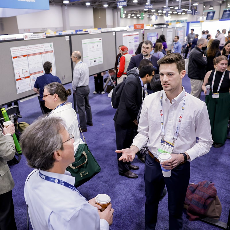 In the foreground, two individuals stand across from each other talking. Around them, you can see others engaged in similar small discussions with poster boards lined up and creating an aisle in which the people stand.