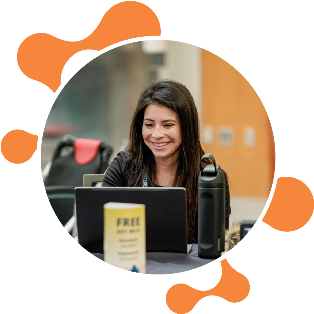 An individual sits at a round table. A laptop and refillable water bottle are sitting on the table in front of them. The individual is smiling, and there are no other individuals visible in the image.