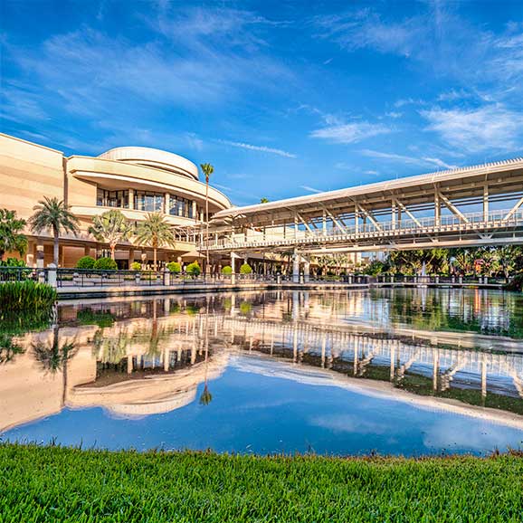A daytime, exterior view of the West Concourse of the Orange County Convention Center in Orlando, Florida. You can see a skybridge leading into the convention center, as well as a pong and landscaping in front of the center.