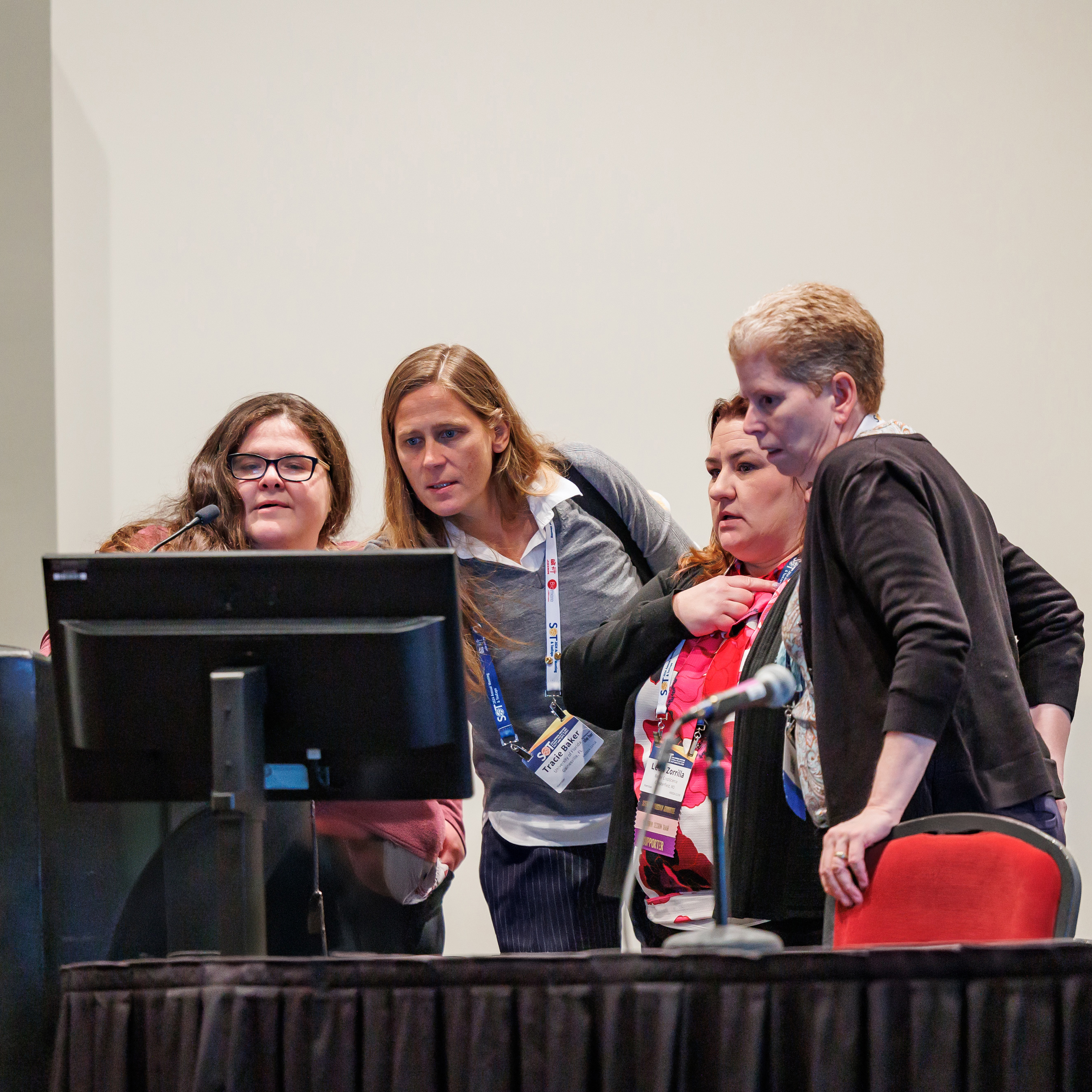 Four individuals stand behind a podium. They are looking at a monitor sitting on the long, skirted table next to the podium.