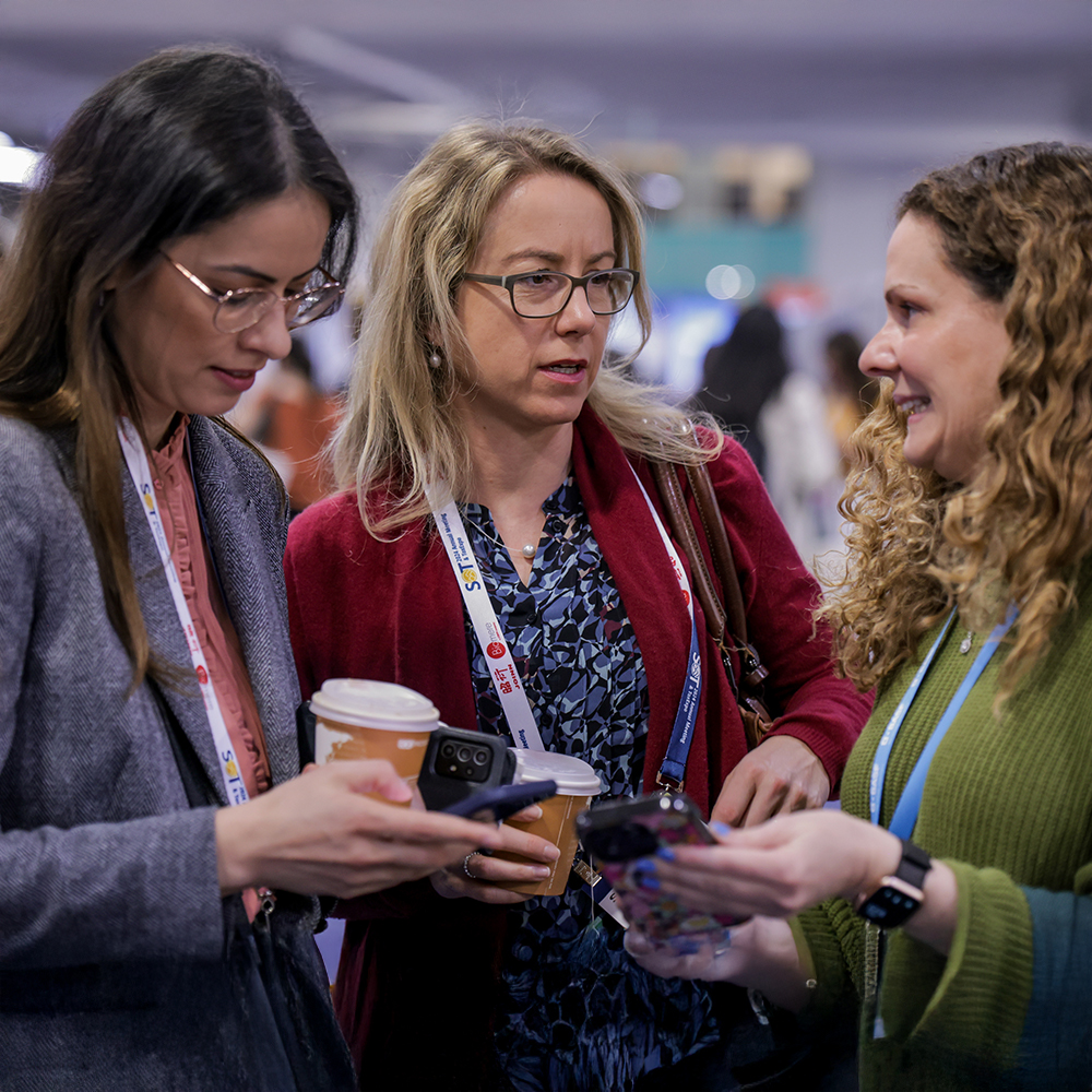 Three individuals stand crowded together with other individuals around them. The three individuals are looking at a phone held by the person in the middle of the small group.