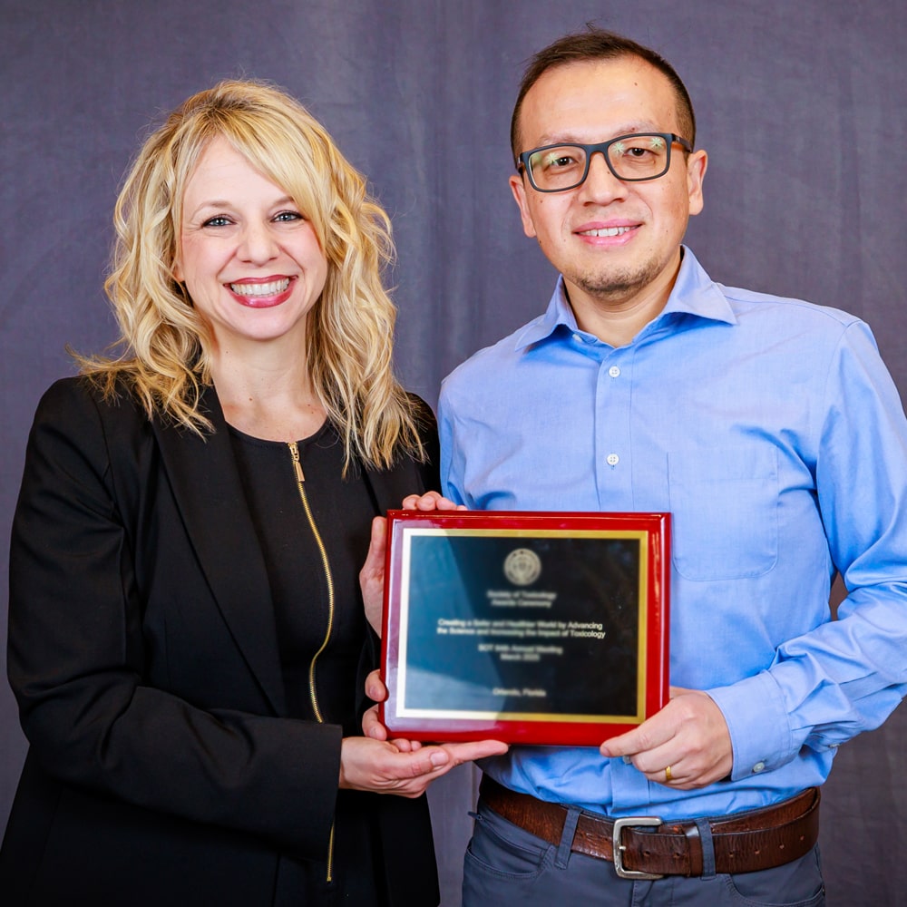 Two individuals stand shoulder-to-shoulder. Between them they are holding an award plaque. Both are looking at the camera and smiling.