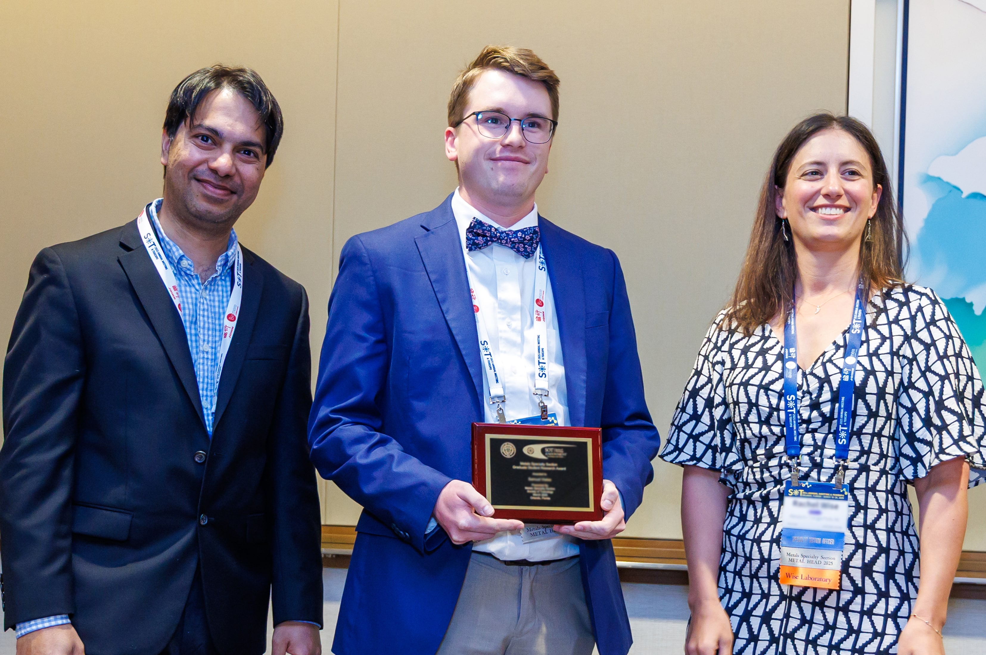 Three individuals stand shoulder-to-shoulder. The individual in the middle is holding an award plaque. All three are smiling.