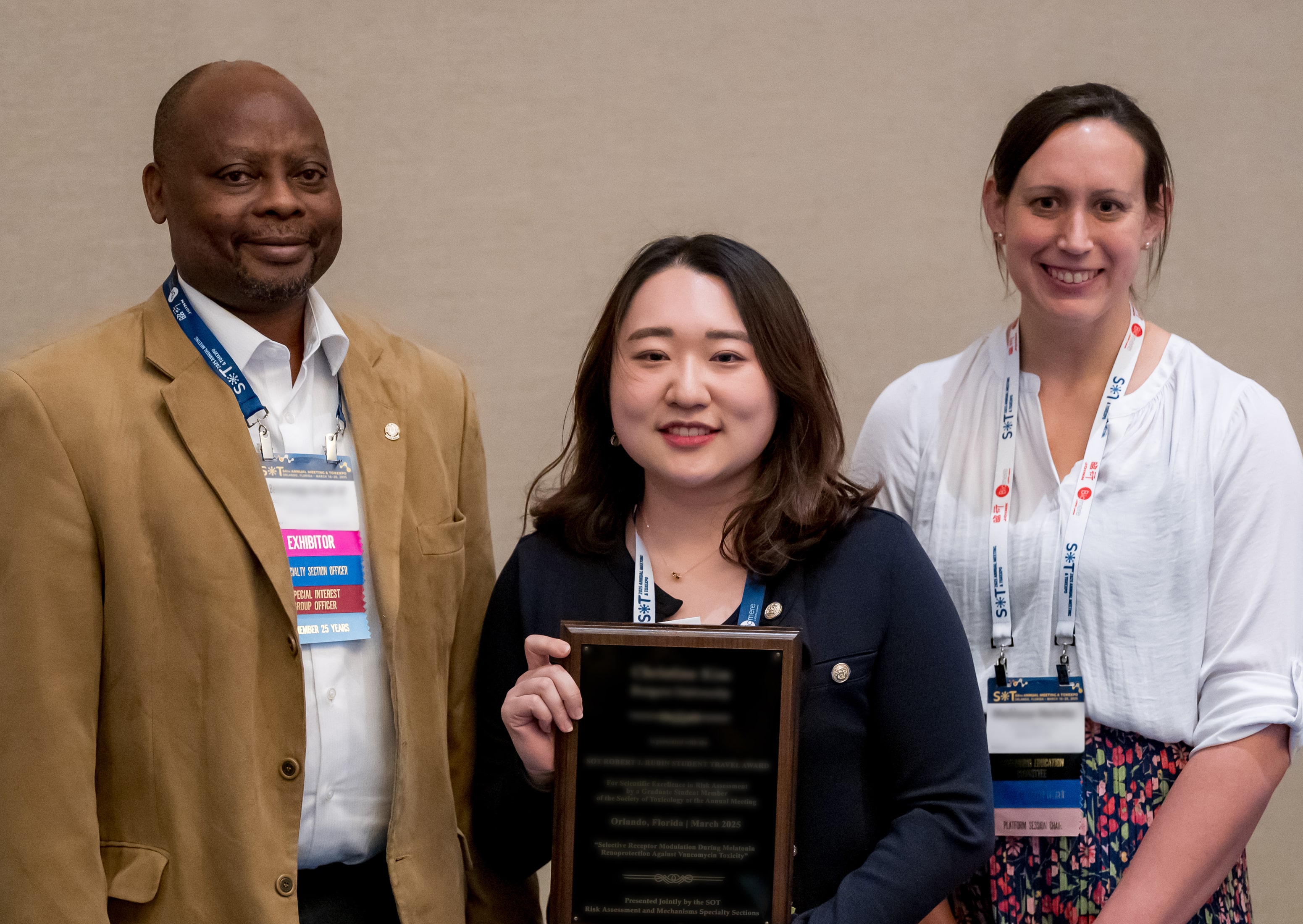 Three individuals stand together. The individual in the middle is holding a large award plaque. All three individuals are smiling at the camera.