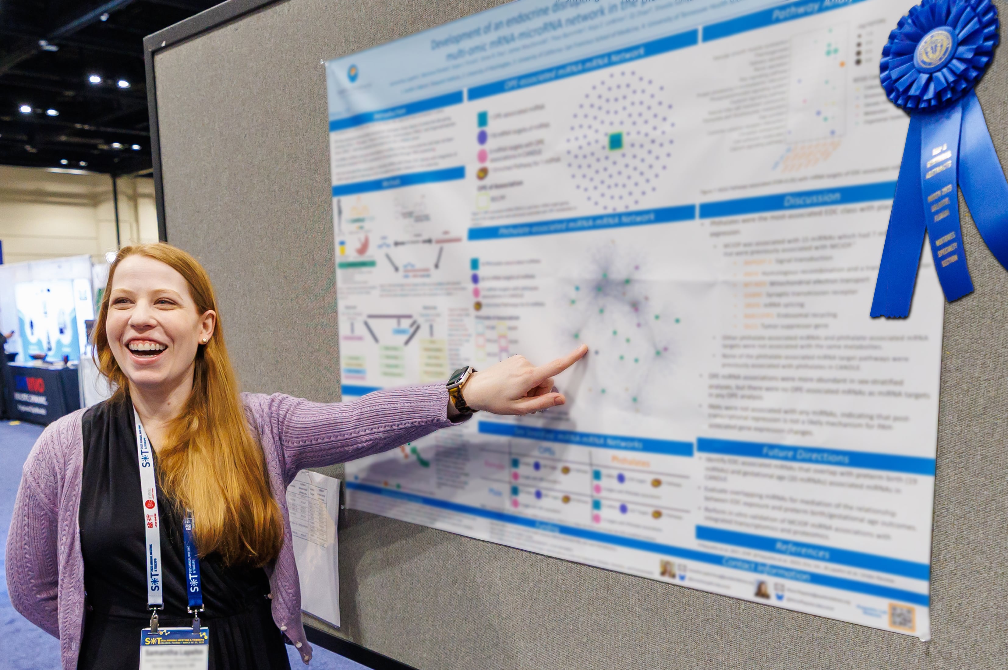 An individual is pointing at a graph on a poster display while smiling at the person to whom they are speaking. Next to the poster, a blue ribbon is displayed.