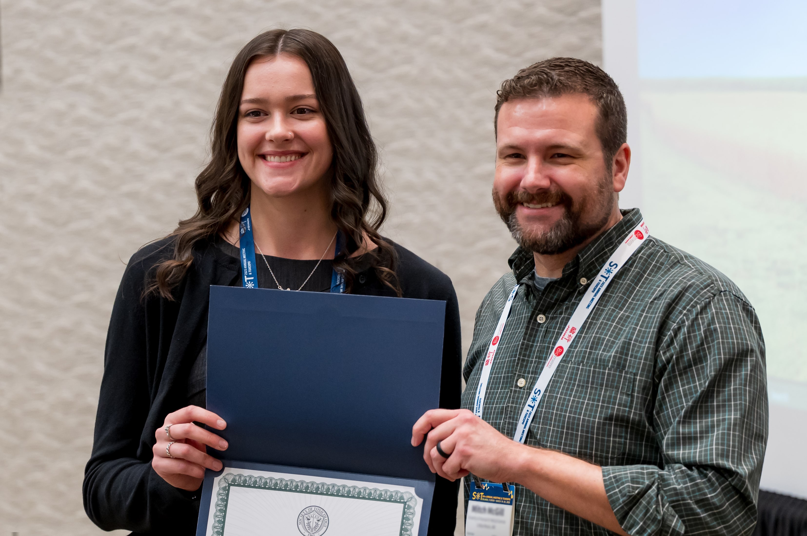 Two individuals are standing shoulder-to-shoulder. Between them, they are holding an award certificate.