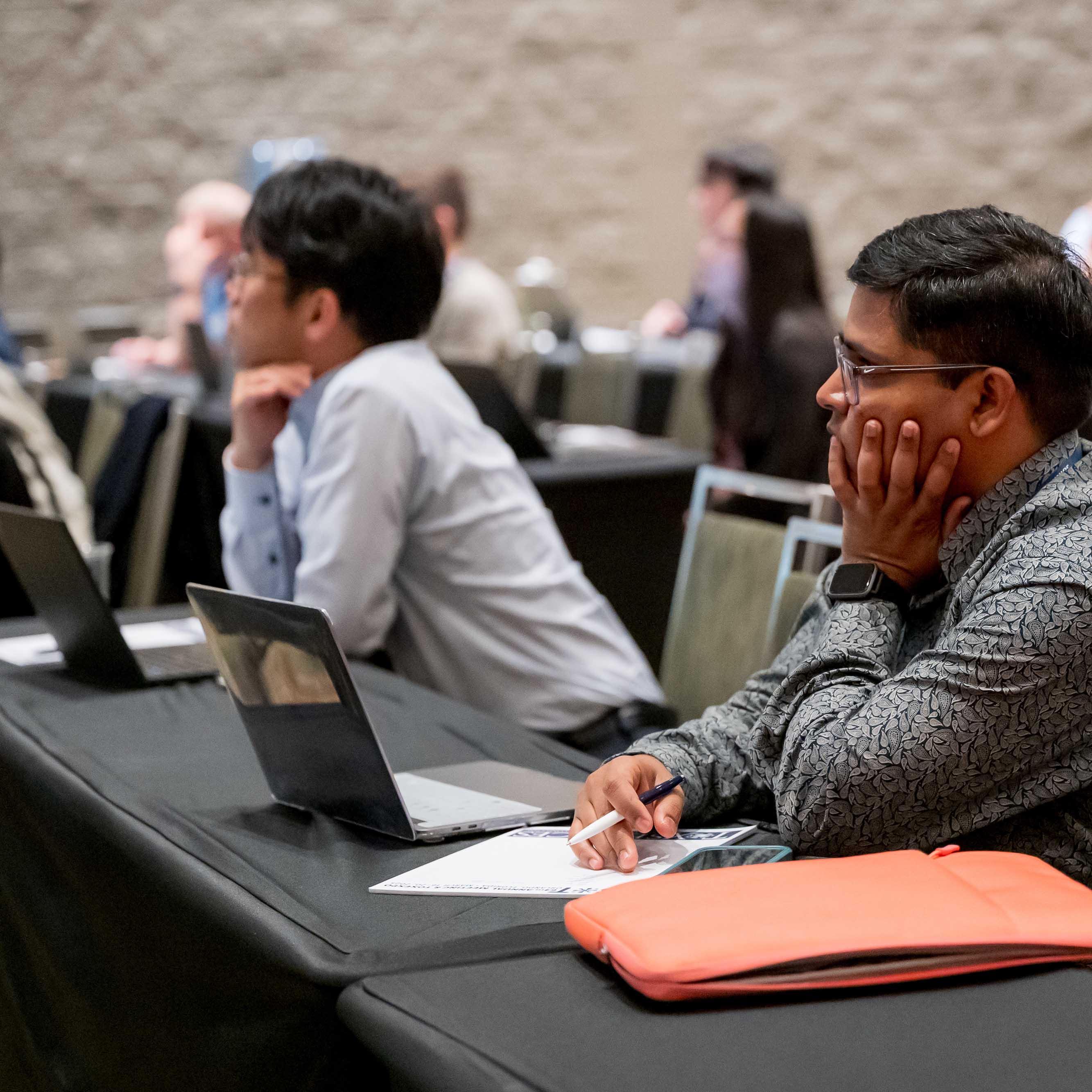 An individual sits at a long table. A laptop sits open on the table. The individual is holding a pen, ready to take notes on a notepad. They are looking toward the front of the room in concentration.
