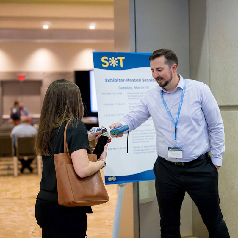  An individual stands next to a sign in front of a doorway. In their hand is an electronic badge scanner. They are in the process of scanning the badge of the individual standing in front of them.