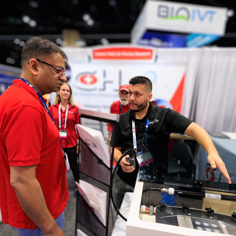 Two individuals are standing near other other. One is leaning over and gesturing at some kind of electronic equipment while the other individual observes.