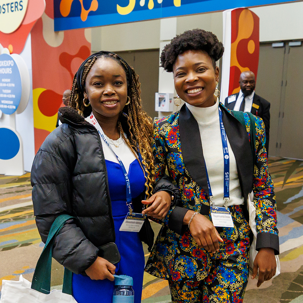 Two individuals stand shoulder-to-shoulder. They have their arms hooked together and are smiling at the camera.