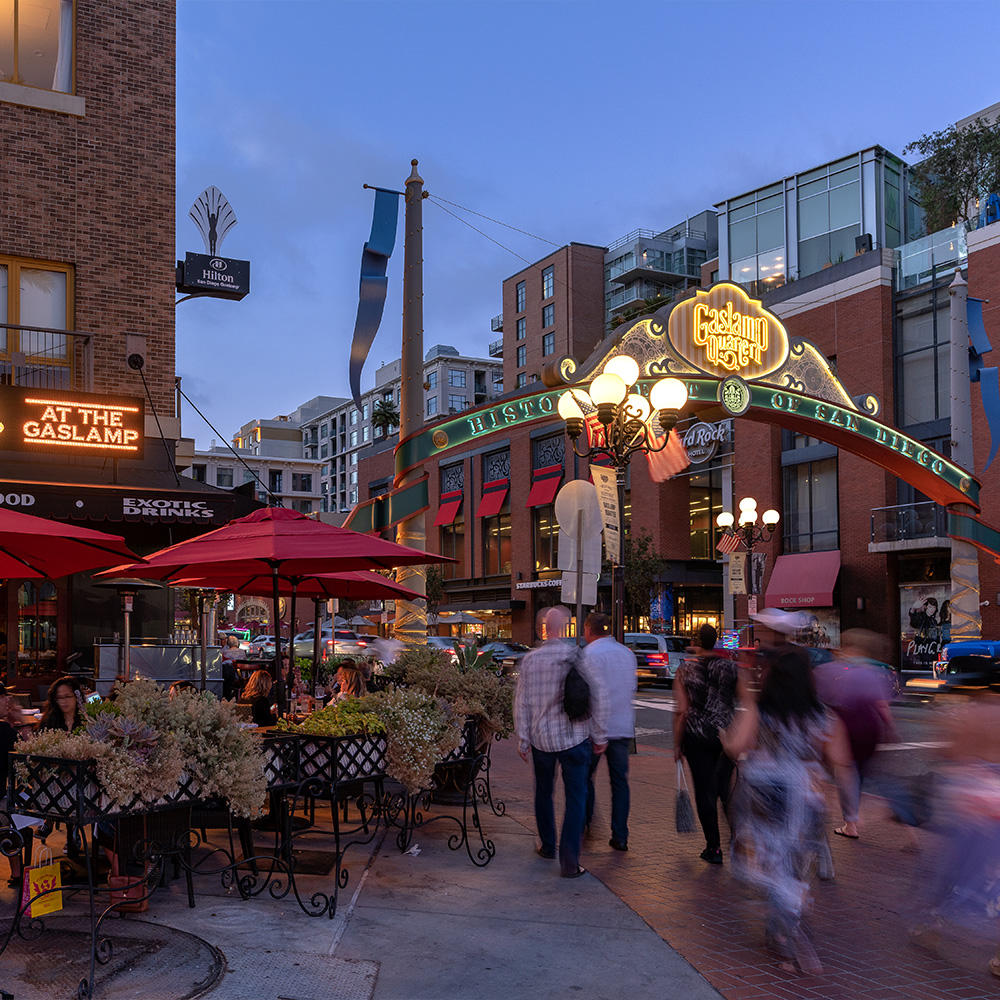 A sign spanning a street reads Gaslamp Quarter. On the corner of this street is a restaurant with people sitting under umbrella tables on a patio. It is nighttime.