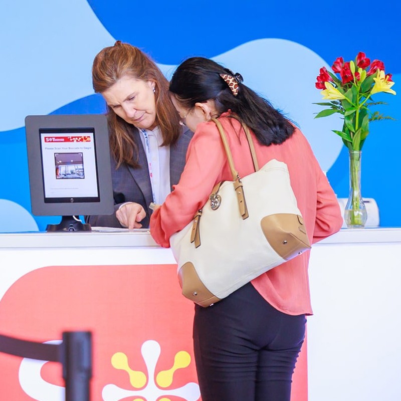An individual stands behind a counter. A person stands across from them on the other side of the counter. Both individuals are looking at something on the counter.